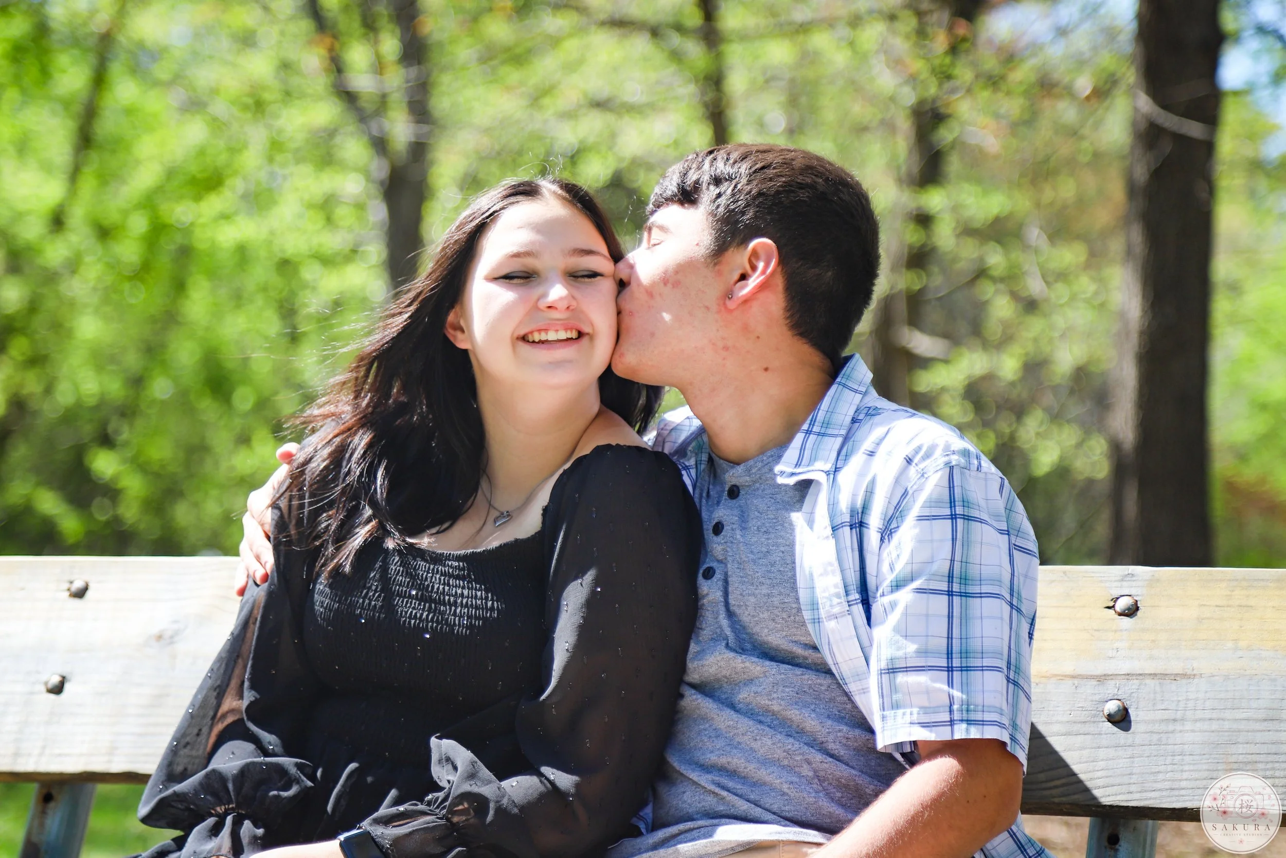 A young man and woman sitting on a bench in a park, the man kissing the woman on the cheek while she smiles with closed eyes. The background is filled with green trees and sunlight.