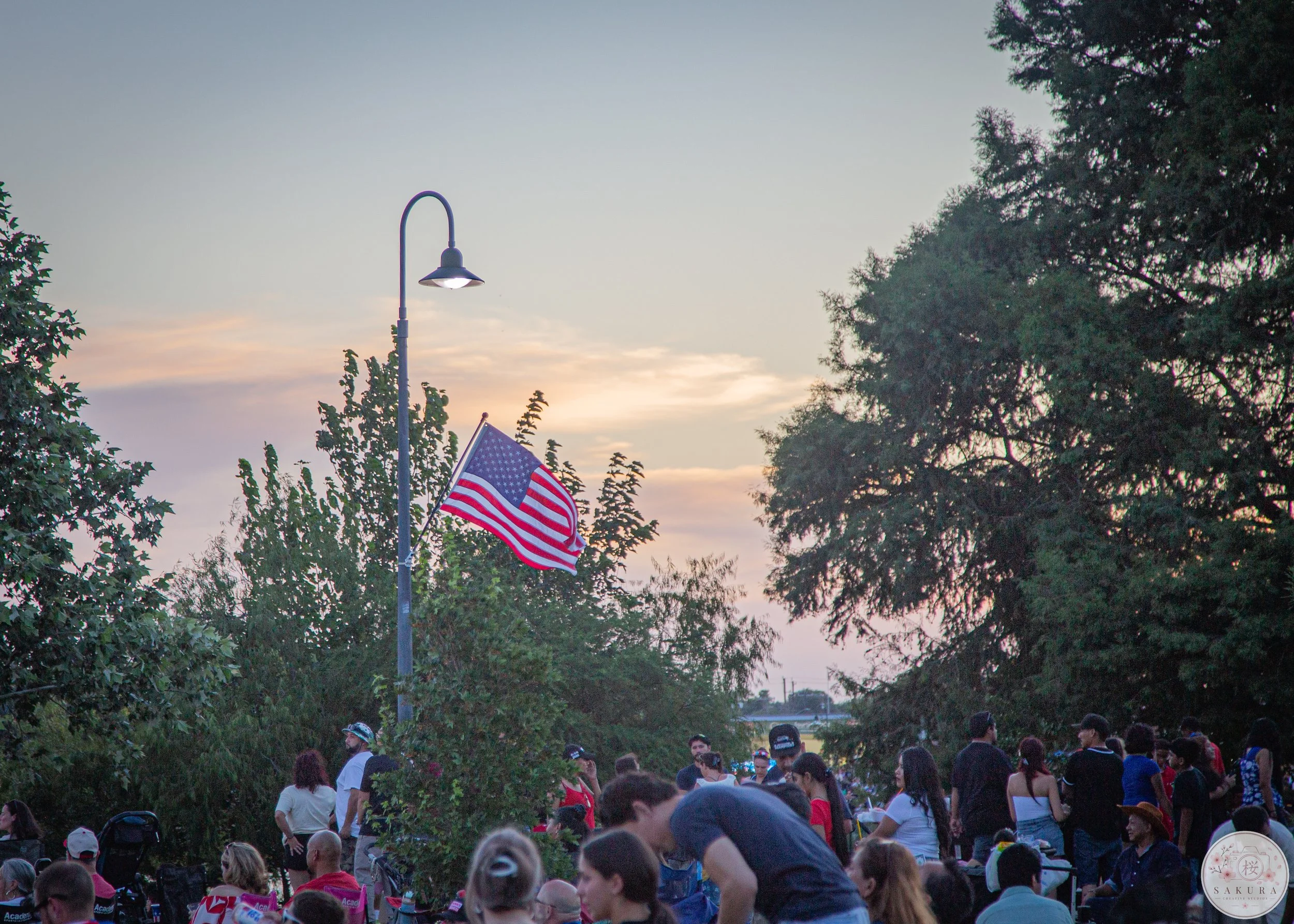 A crowd of people gathered outdoors at sunset, with an American flag flying and trees surrounding the area.