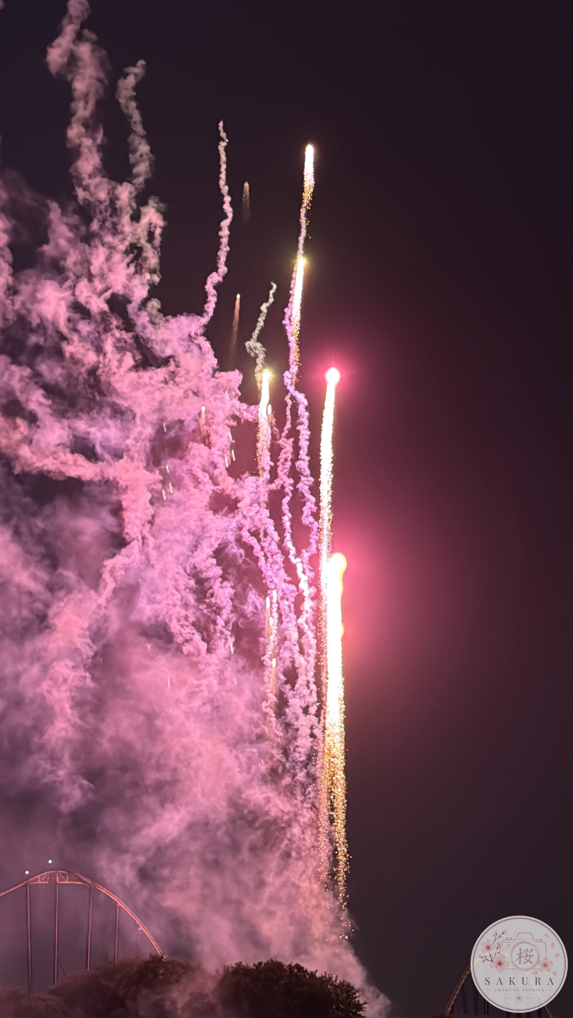 Fireworks illuminating the night sky with pink and white colors, creating trails and smoke, as part of a celebration event.