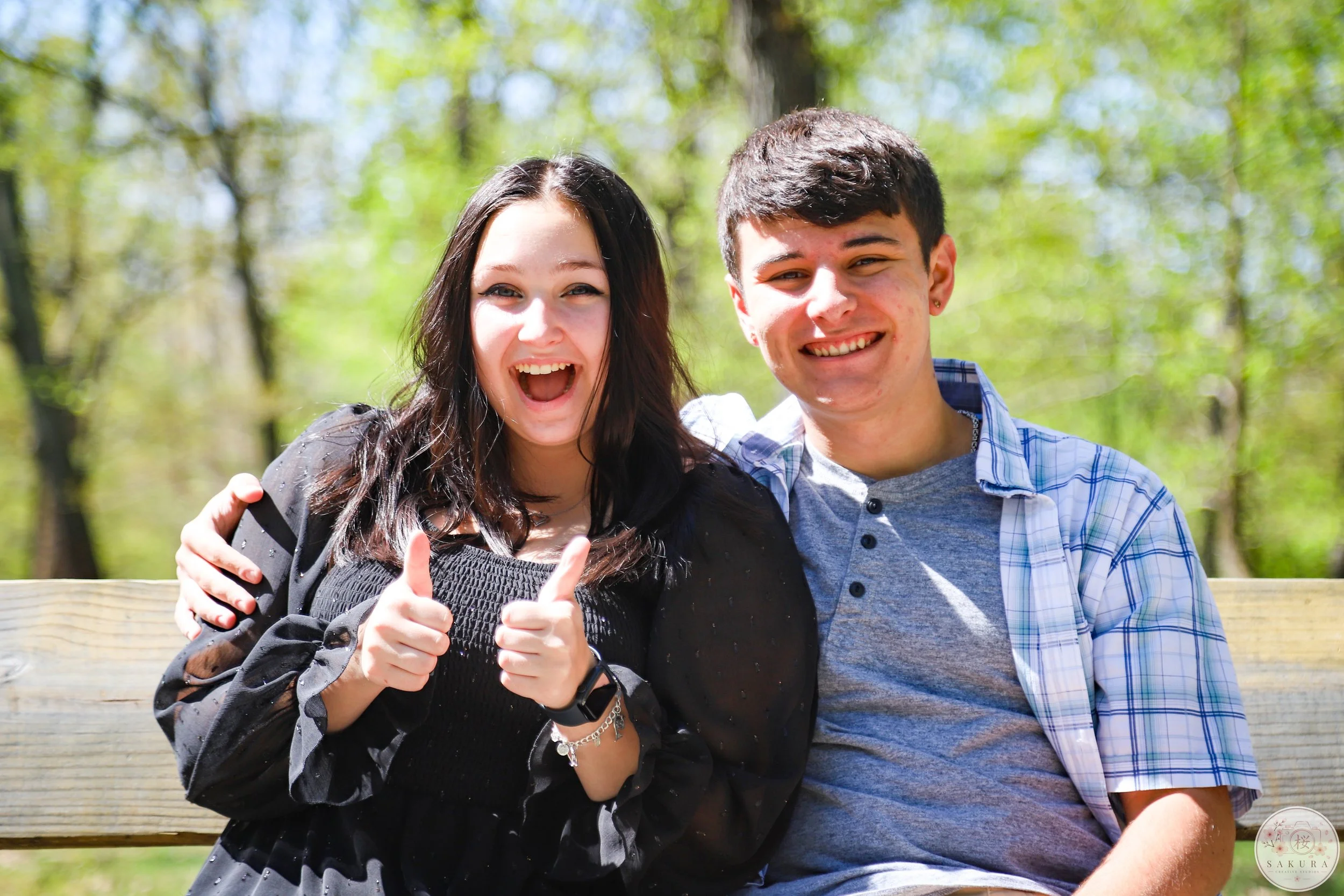 A young woman and young man are sitting closely together on a wooden bench outdoors in a park with green trees in the background. The woman has long dark hair, is smiling widely with her mouth open, and is giving a thumbs-up. The man has short dark h