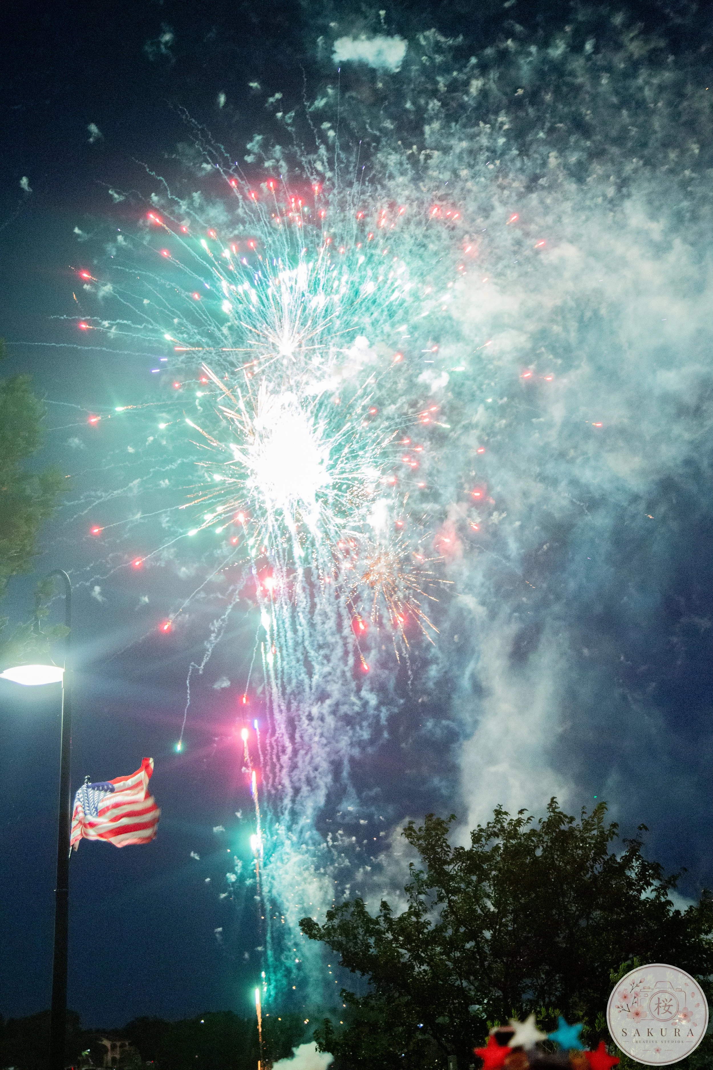 Colorful fireworks light up the night sky during celebration, with an American flag on a flagpole visible on the left side and some trees in the foreground. The image also features a logo with cherry blossom and Japanese characters at the bottom righ