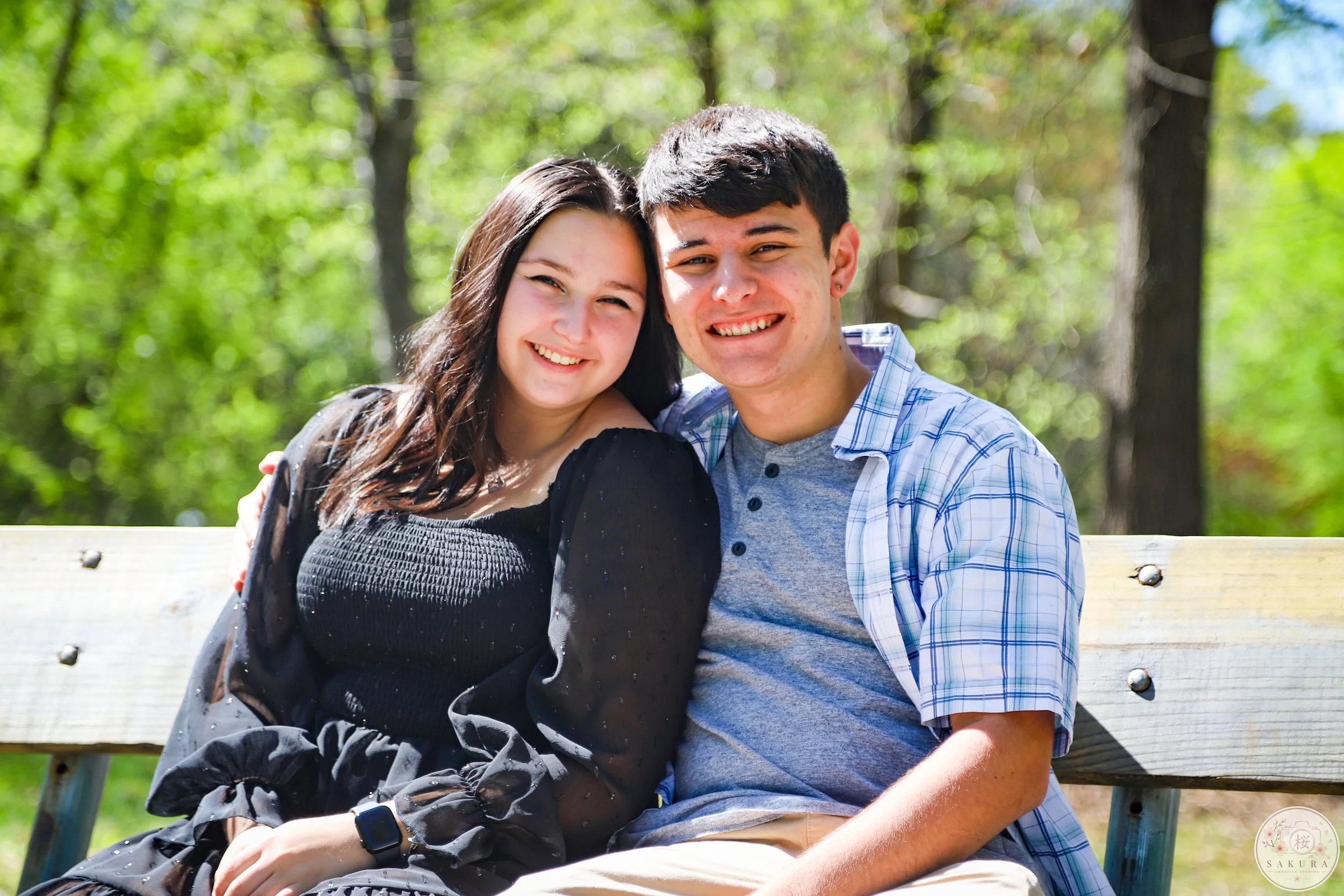 A young man and woman sitting closely on a wooden bench outdoors, smiling at the camera, surrounded by green trees.