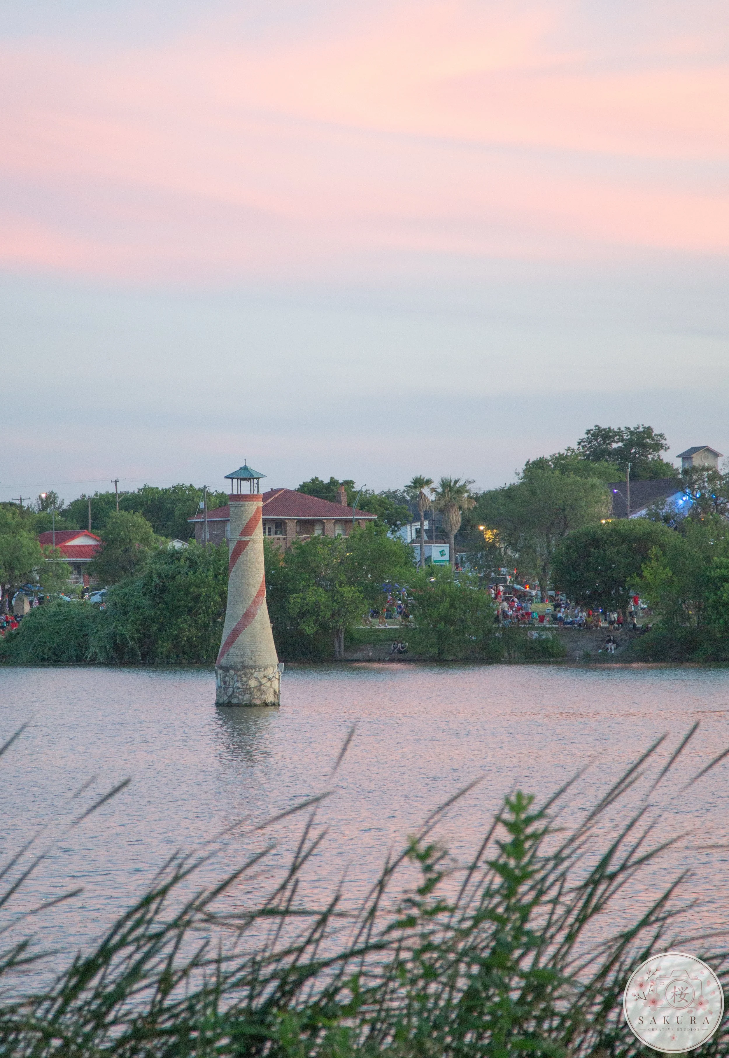 A lighthouse next to a body of water with houses and trees along the shoreline, and a pastel-colored sky at dusk.