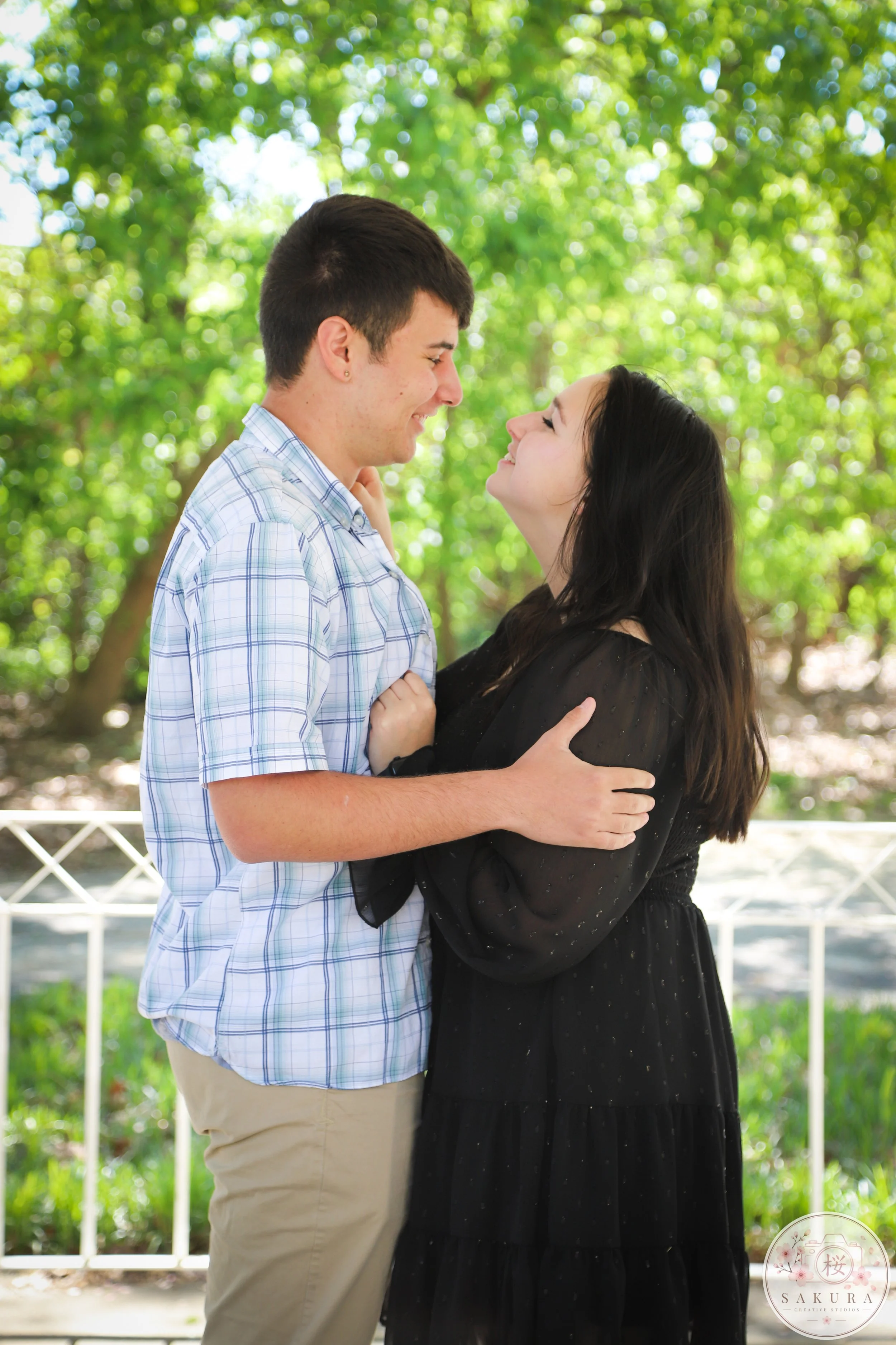 A young couple standing close together outdoors, smiling and looking into each other's eyes, surrounded by green trees.