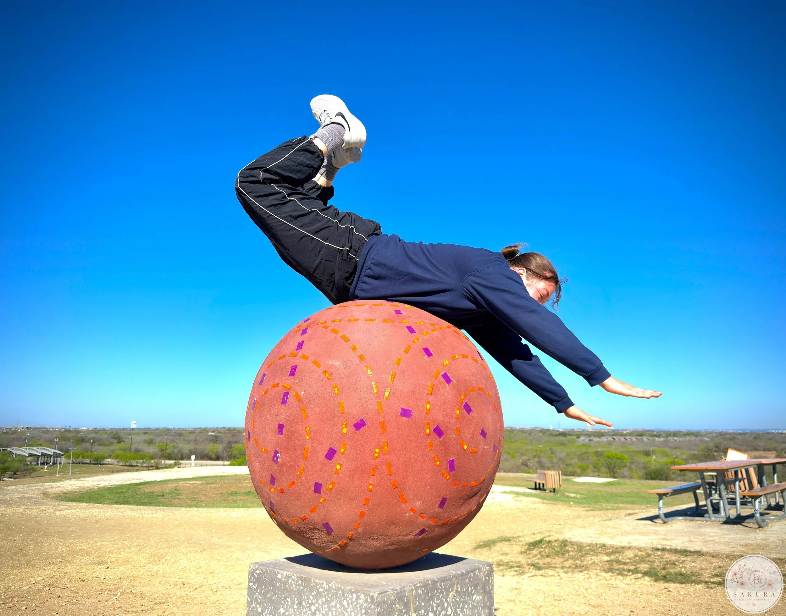 Person performing a yoga pose, lying on a large decorative sphere outdoors against a clear blue sky.