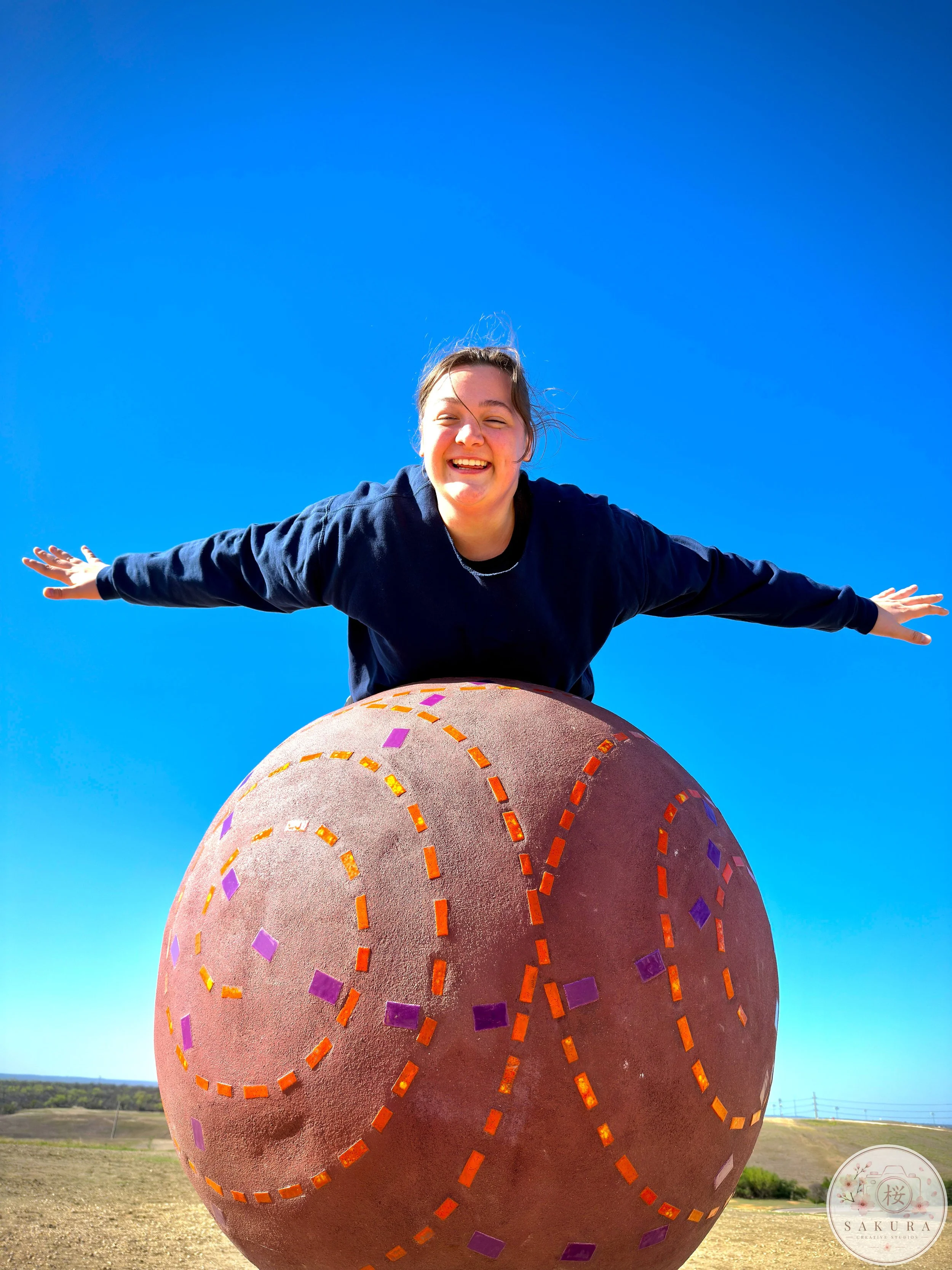 A young woman with brown hair, wearing a navy sweatshirt, smiling and spreading her arms wide while sitting on a large, decorated sphere outdoors under a bright blue sky.