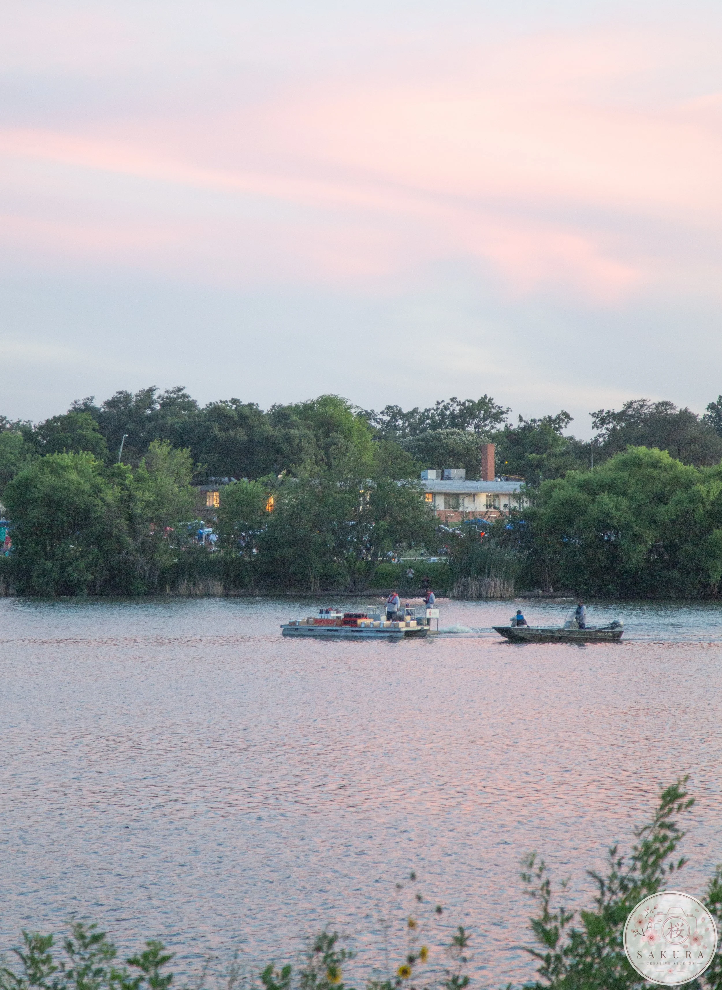 Two boats floating on a calm river with greenery on the banks and houses in the background. The sky is pink and purple, indicating early evening or sunset.