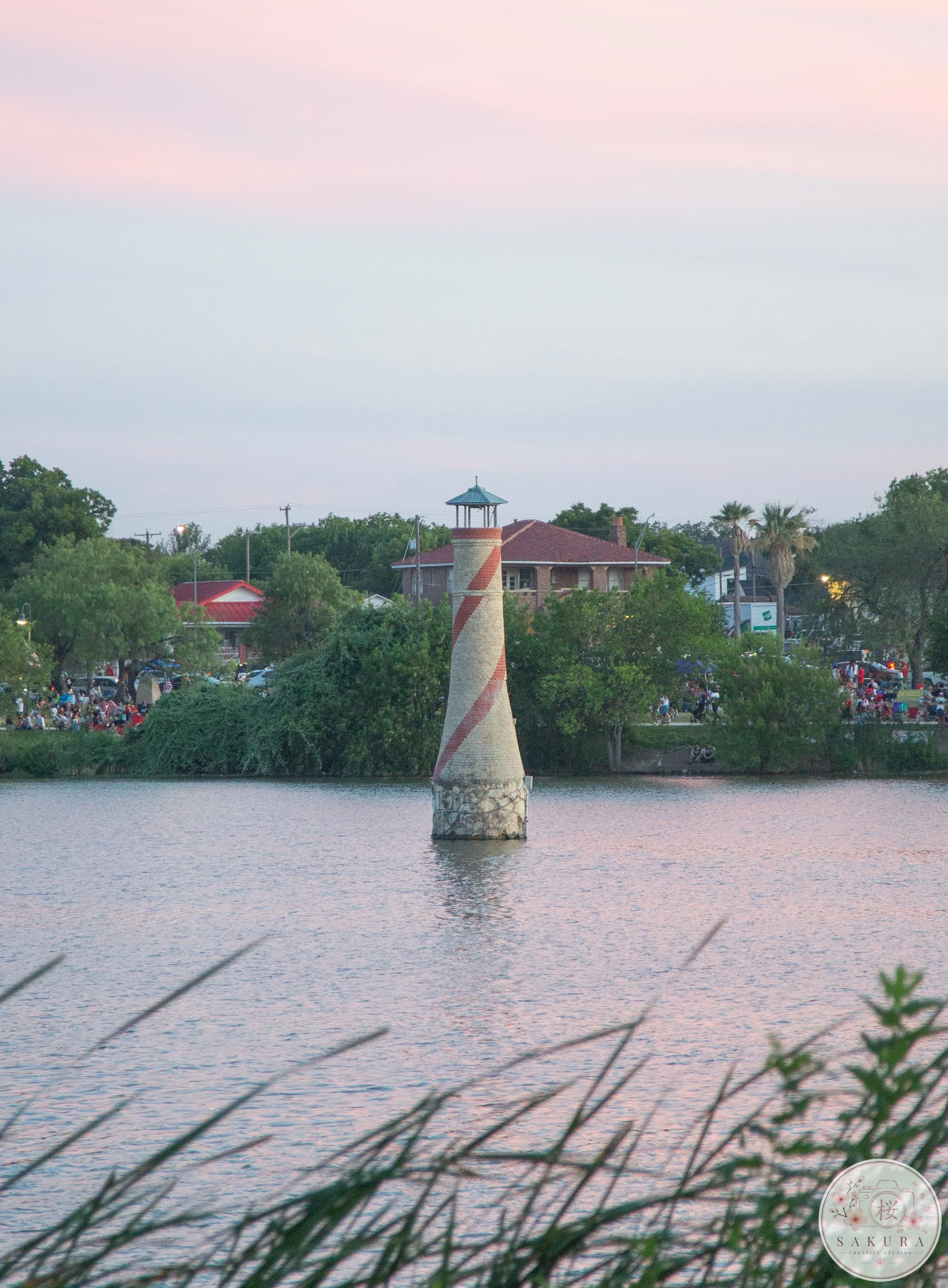A lighthouse on the water with buildings and trees in the background under a pink and blue sky at dusk.