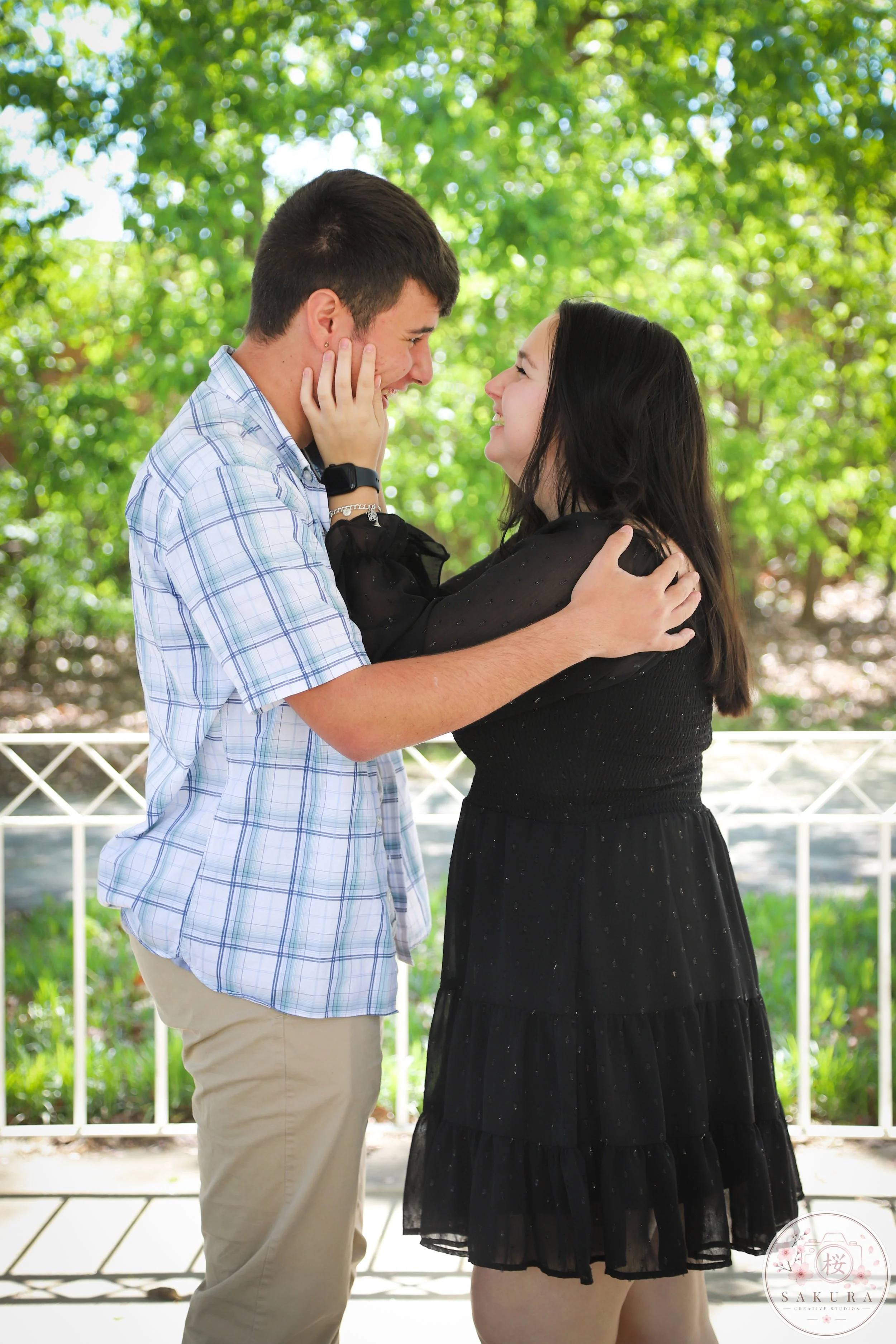 A young couple is standing outdoors on a sunny day, looking into each other's eyes and smiling, with a green leafy background and a white railing.