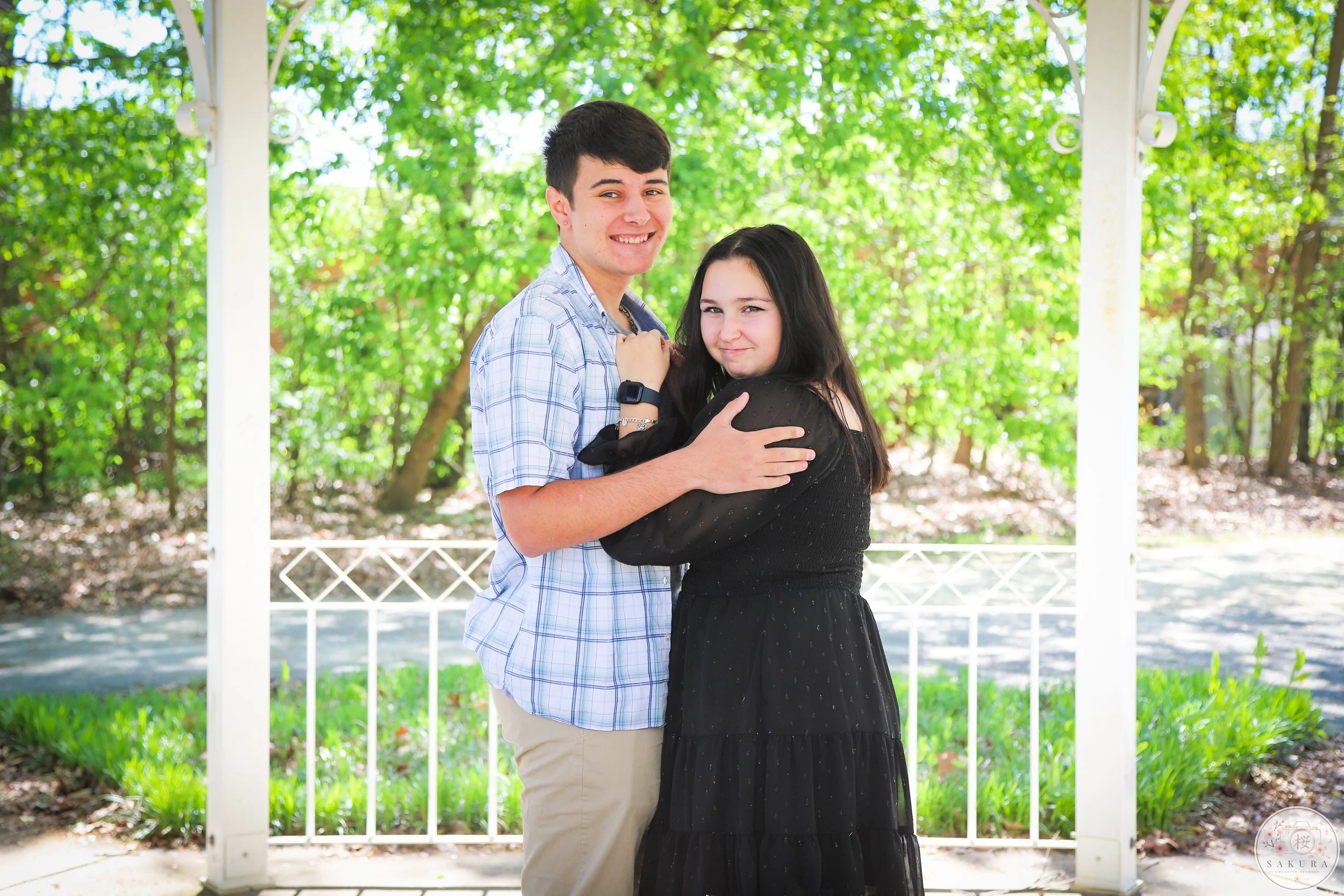 A young man and woman are embracing each other and smiling outdoors on a sunny day with green trees in the background.