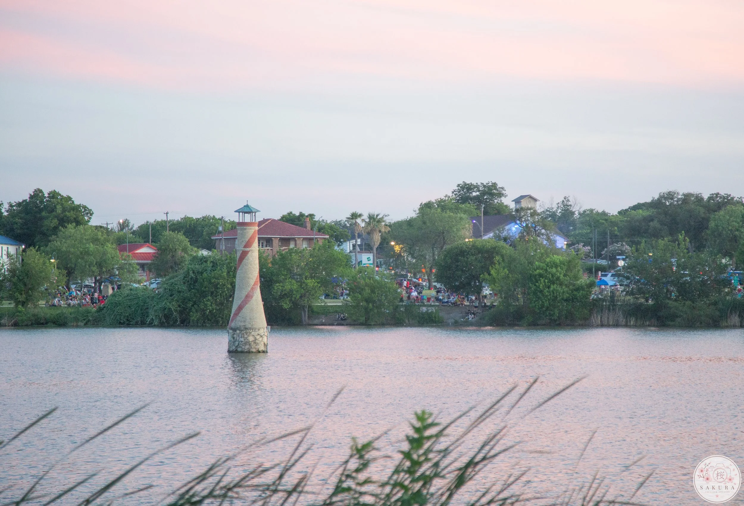 A lake with a small lighthouse in the water, surrounded by trees and buildings in the distance during sunset or evening.
