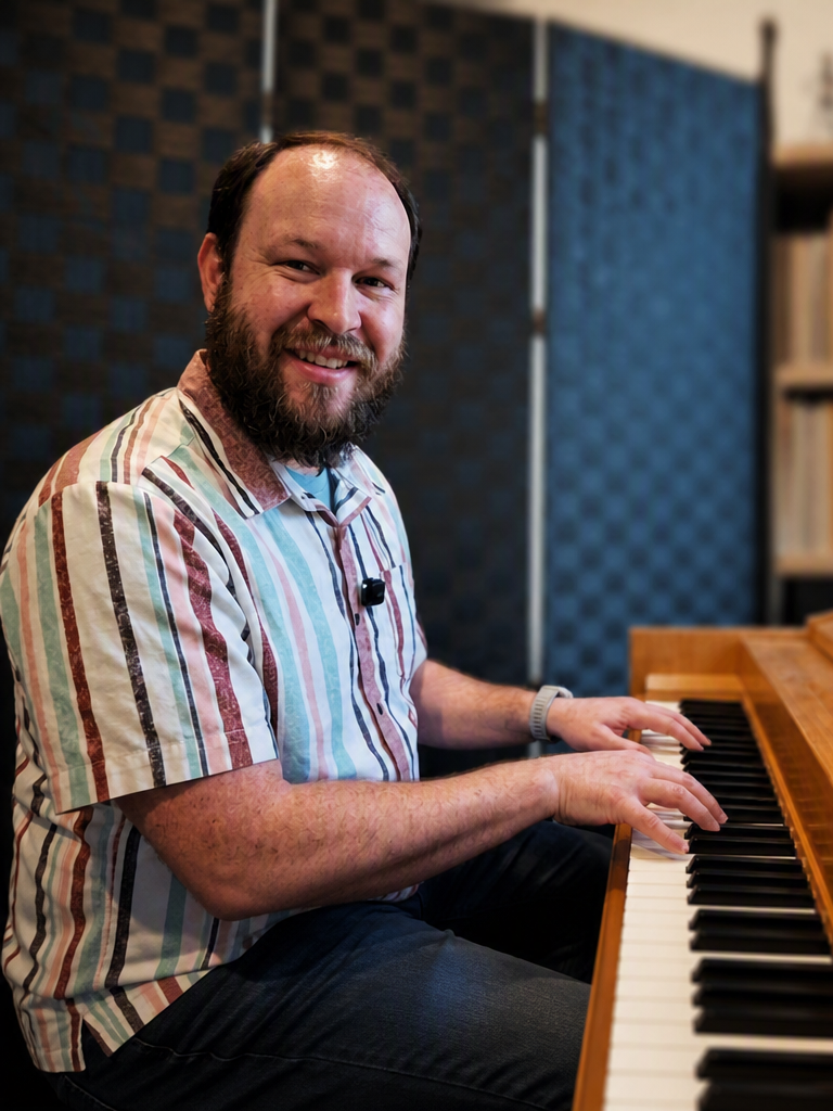 A smiling man with a beard playing a wooden piano in a music studio.