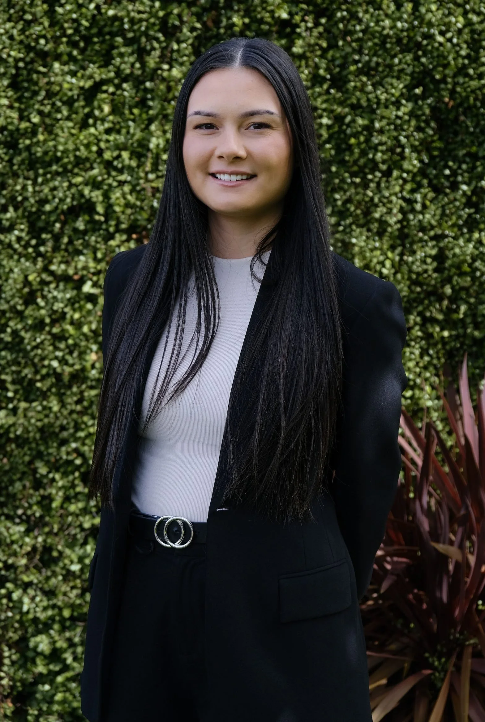 Young woman with long black hair, smiling, wearing a black blazer, white top, and black pants with a silver belt, standing outdoors in front of green foliage.