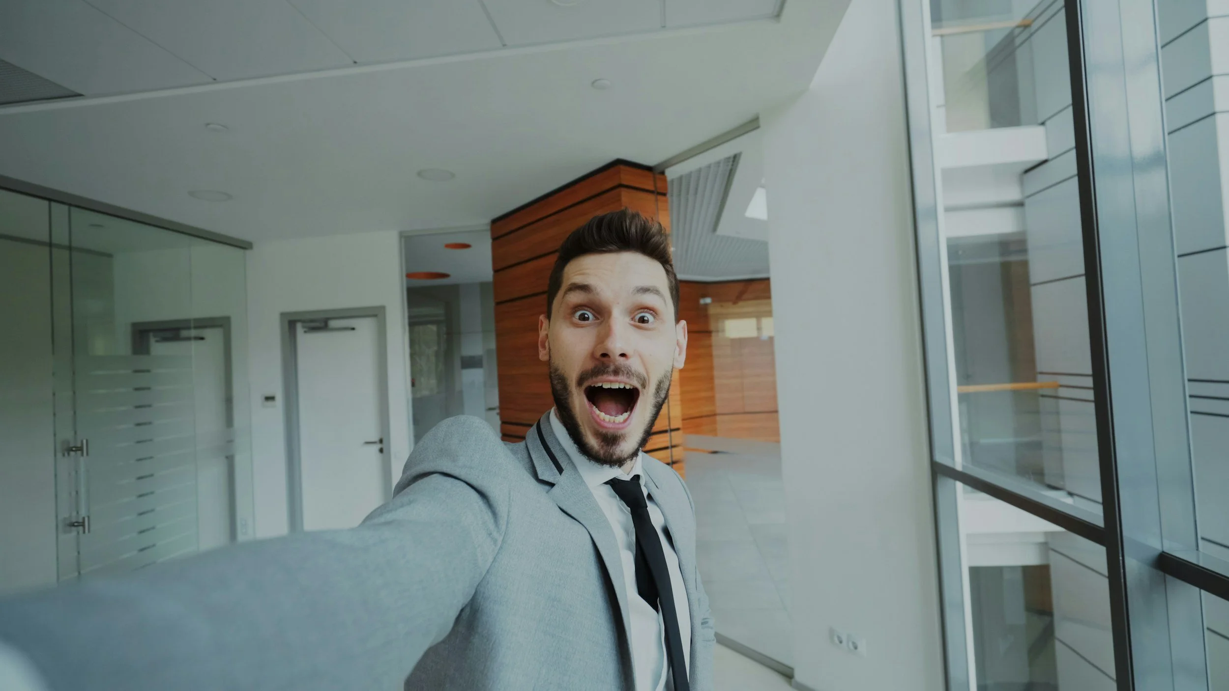 A man in a gray suit taking a selfie inside a modern building with glass walls, wooden accents, and white ceilings, smiling and appearing excited.