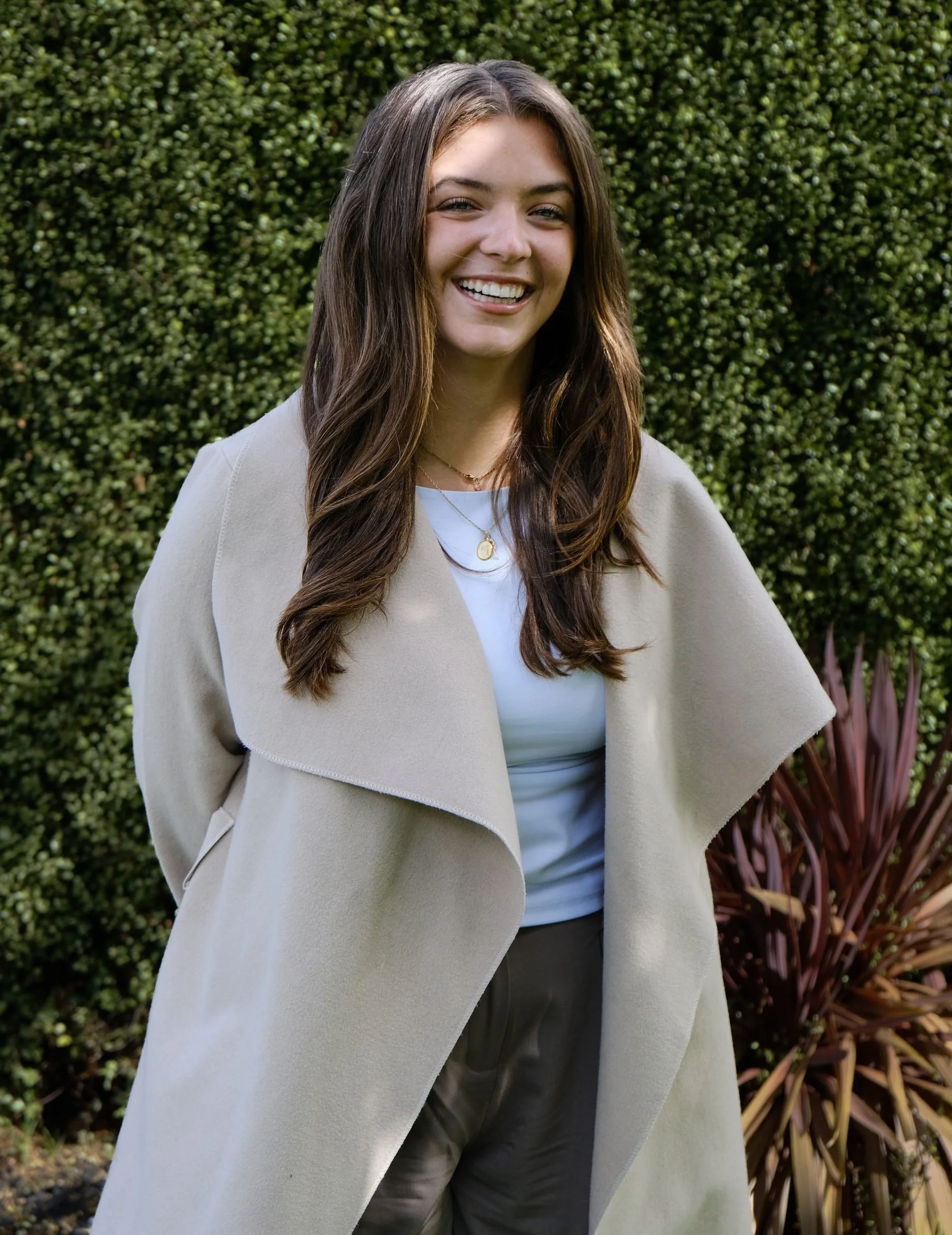 A smiling woman with long brown hair wearing a light beige coat, white shirt, and layered necklaces, standing outdoors in front of a green hedge and plants.