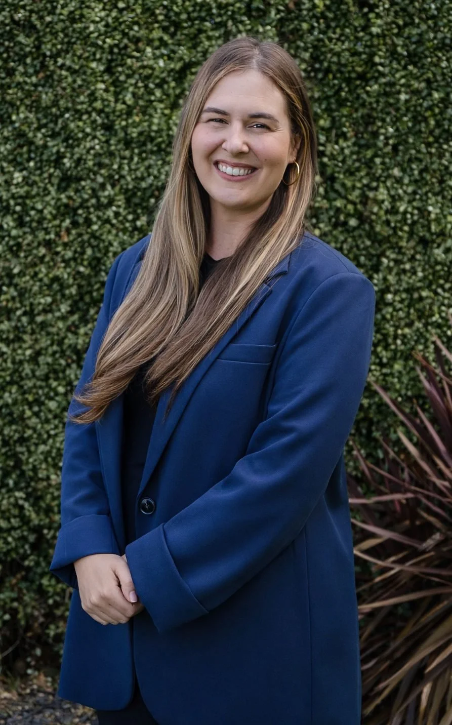 A woman with long light brown hair, wearing a blue blazer and black top, smiling outdoors with green bushes in the background.