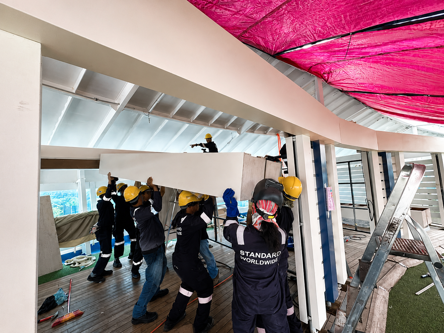 Construction workers wearing yellow helmets and dark uniforms installing a large white panel on a building's structure. One worker is on an upper level, pointing, while others are lifting and positioning the panel below. There is a ladder on the right side, and construction tools and materials are scattered on the floor.