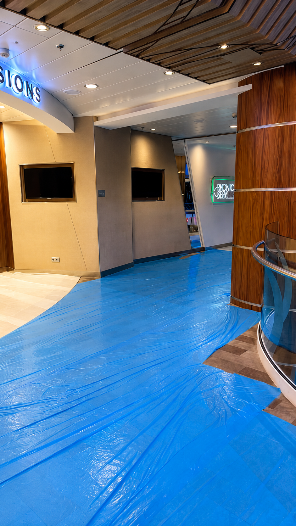 Interior of a modern building with a curved blue plastic sheet covering the floor, mounted TV screens on the wall, wooden panels, and a staircase with glass and metal railing. Neon signs are visible in the background.