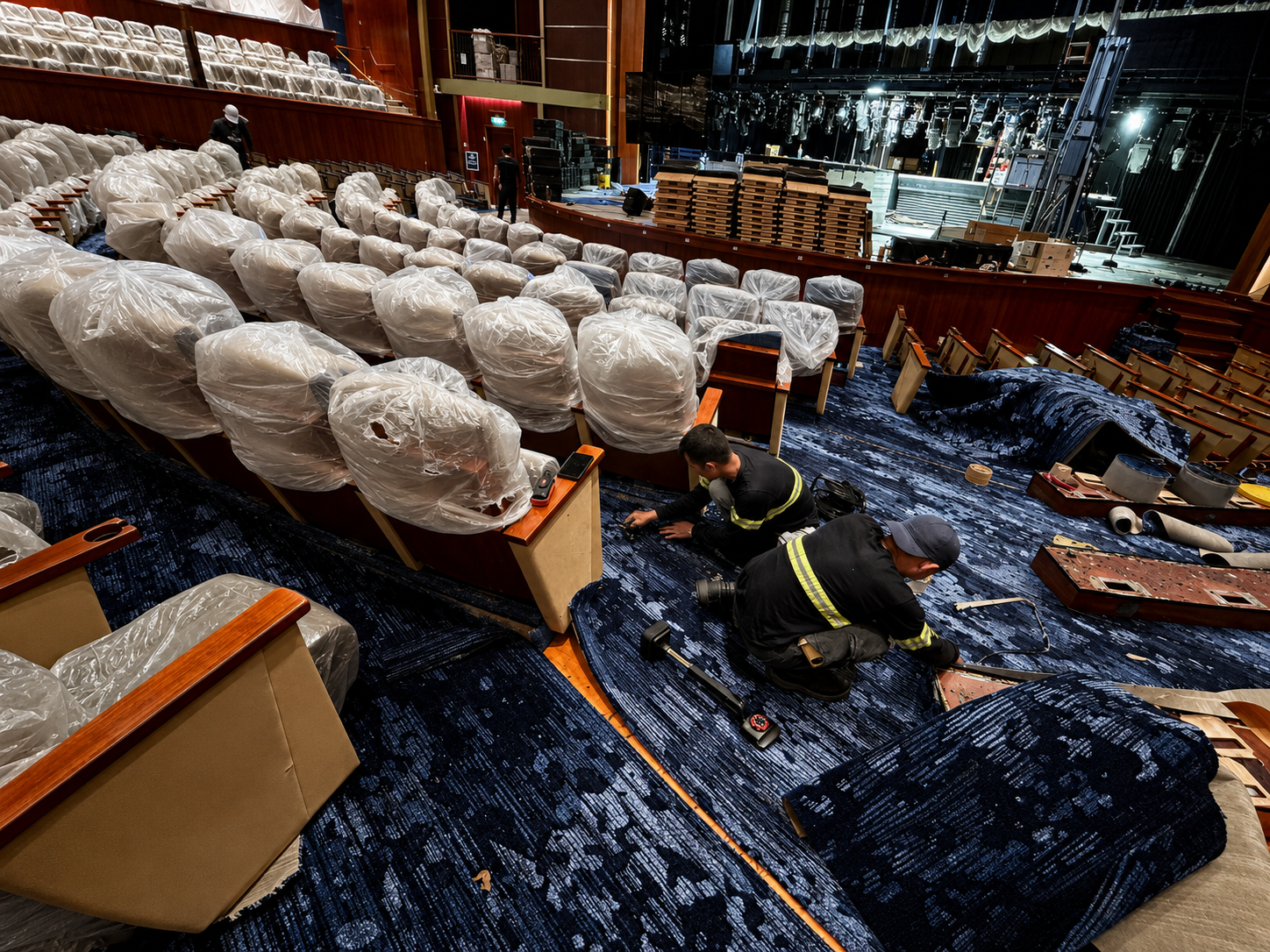Workers installing or repairing blue carpet in an auditorium or theater, with chairs wrapped in plastic and a stage with equipment in the background.