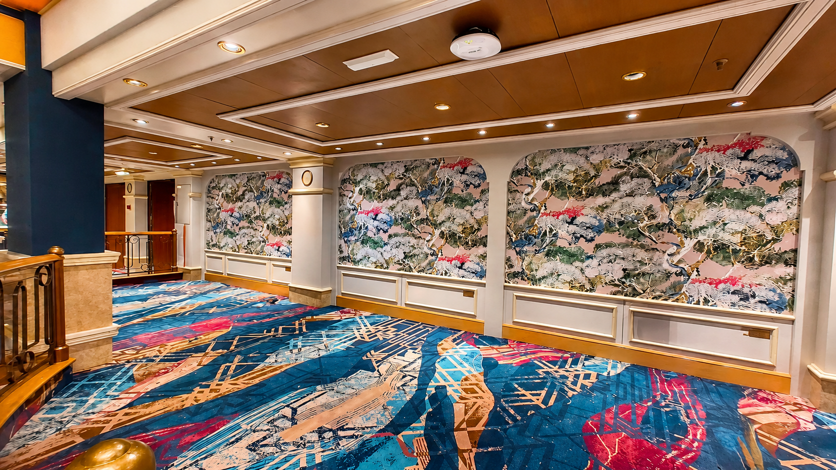 Interior view of a hotel corridor with colorful abstract carpet, decorative wall panels featuring a nature-inspired design, wooden ceiling panels with recessed lighting, and a railing with gold accents.