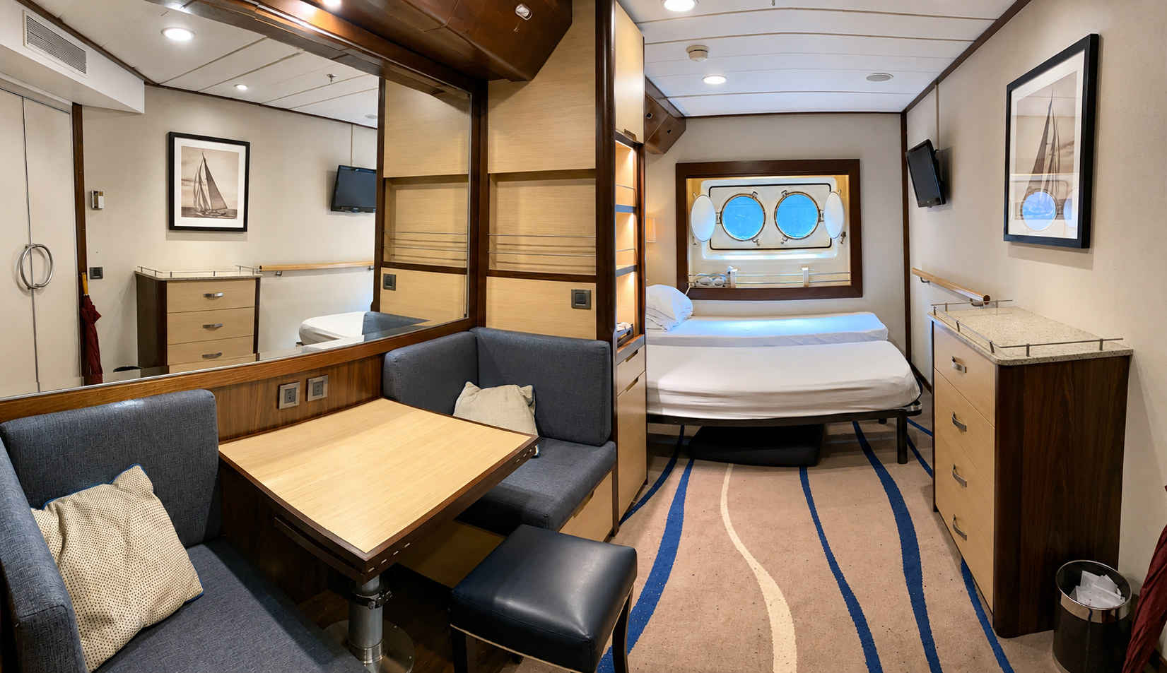 Interior of a cruise ship cabin with a bed, seating area, TV, and portholes showing blue sky.