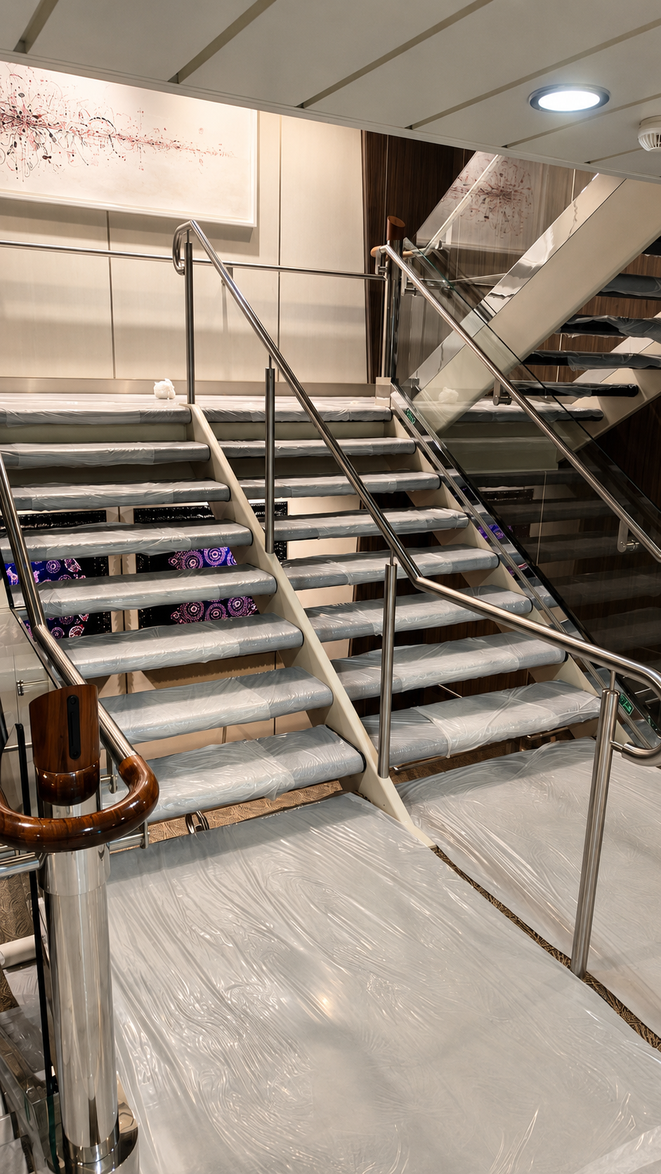 Indoor staircase with metal handrails and plastic-wrapped steps, leading to a higher floor with glass railing and artwork on the wall.