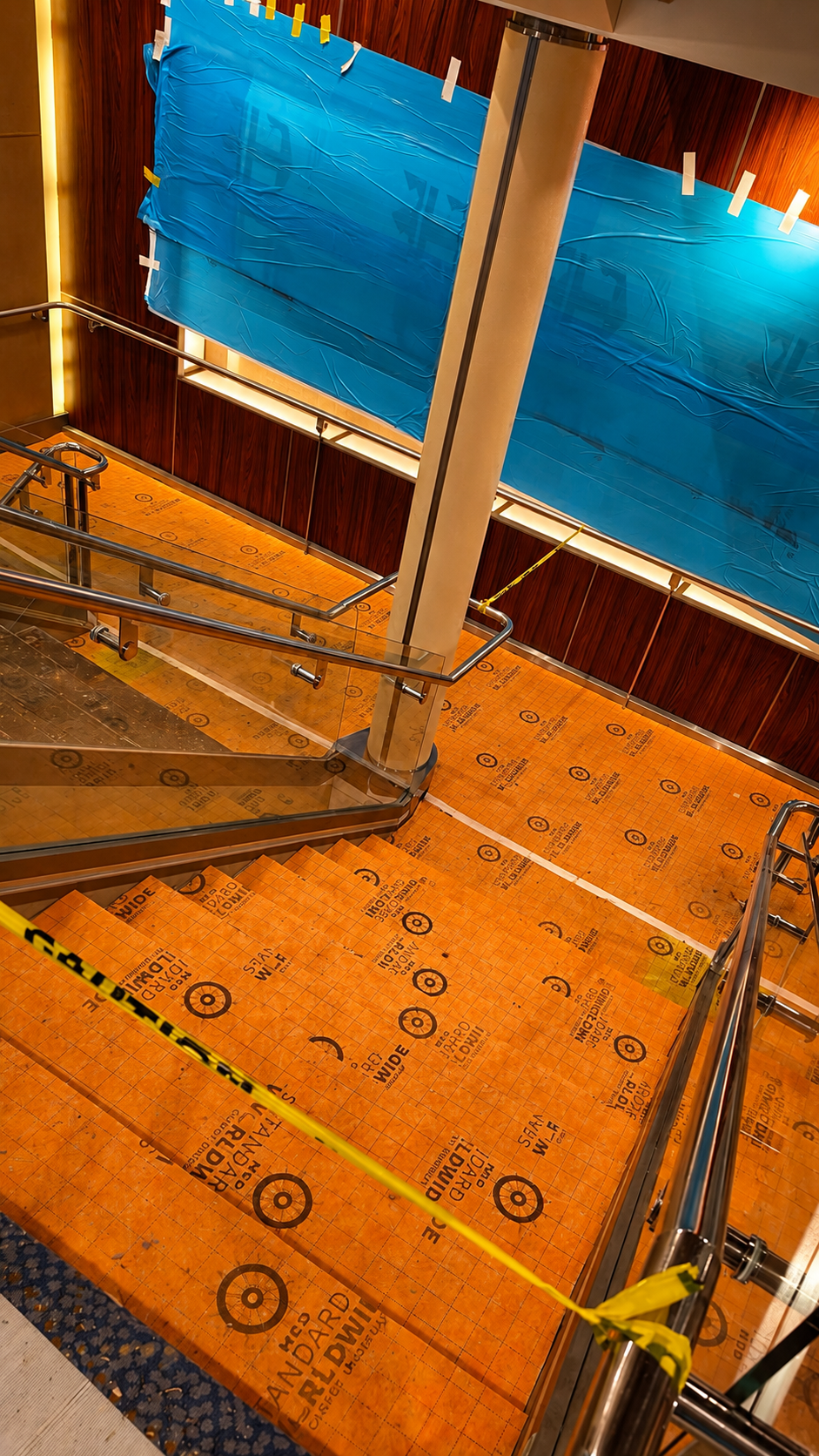 Indoor staircase surrounded by wood-paneled walls, with a large blue plastic sheet taped to the wall, and orange floor protection covering the steps, marked with warning symbols.