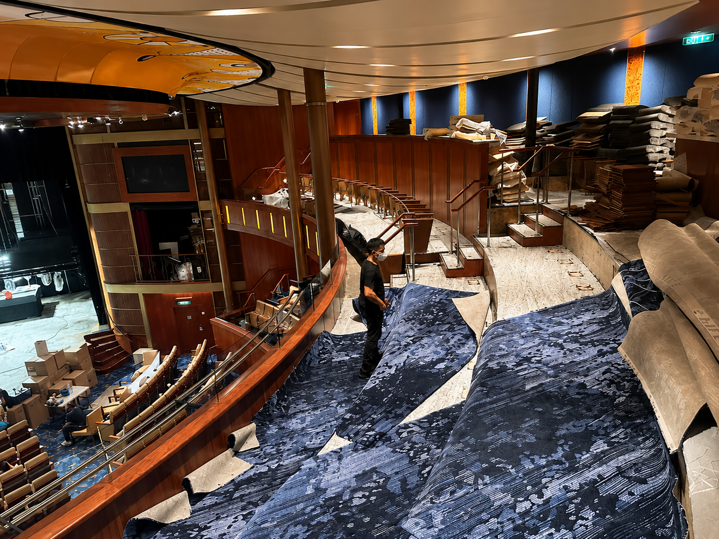 Interior of a theater or auditorium under renovation with a person standing on a partially covered stage, stacks of fabric or carpet rolls, and seating area visible below.