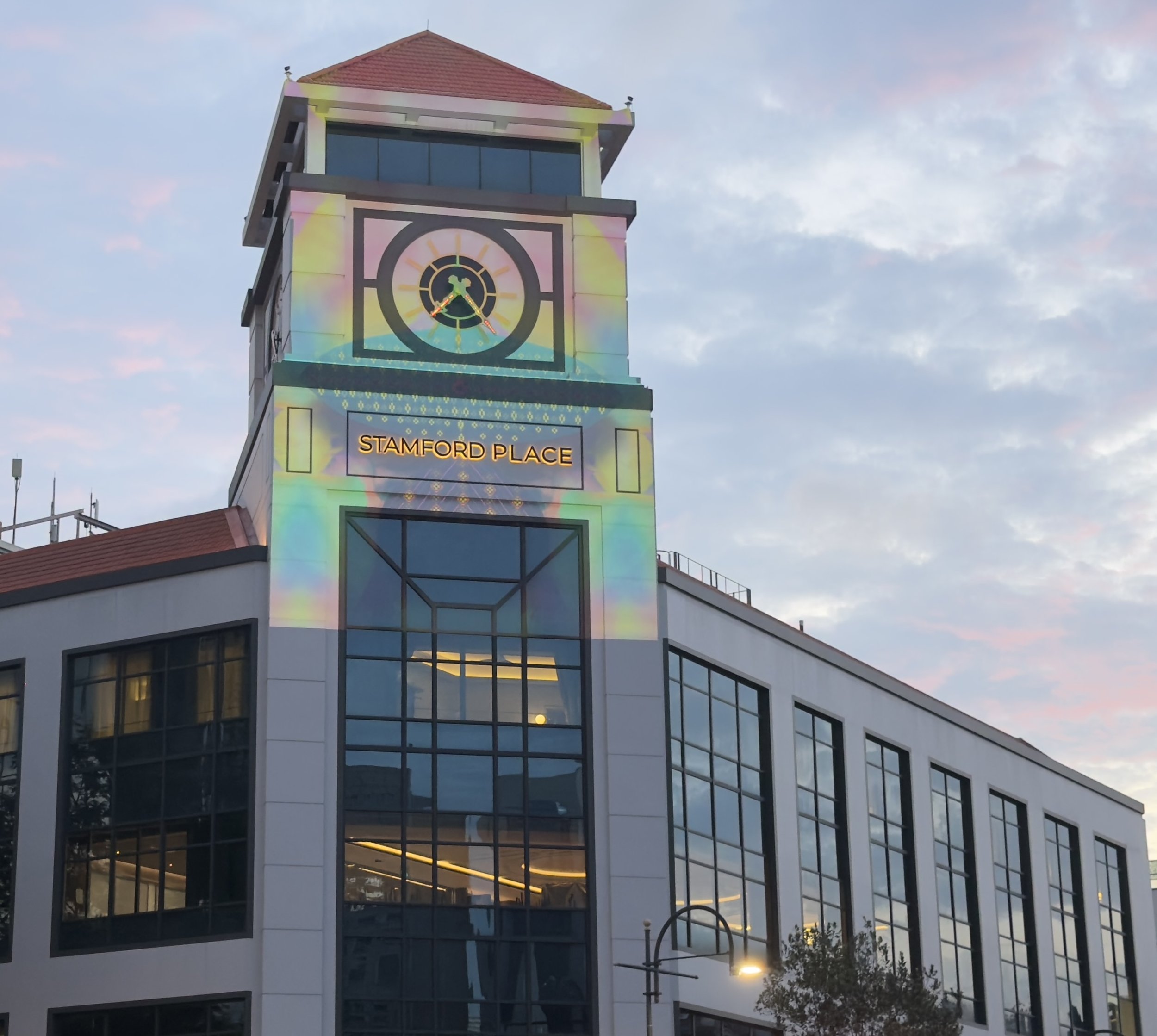 Clock tower building with colorful light projection, labeled 'Stamford Place,' sky with clouds at dusk.
