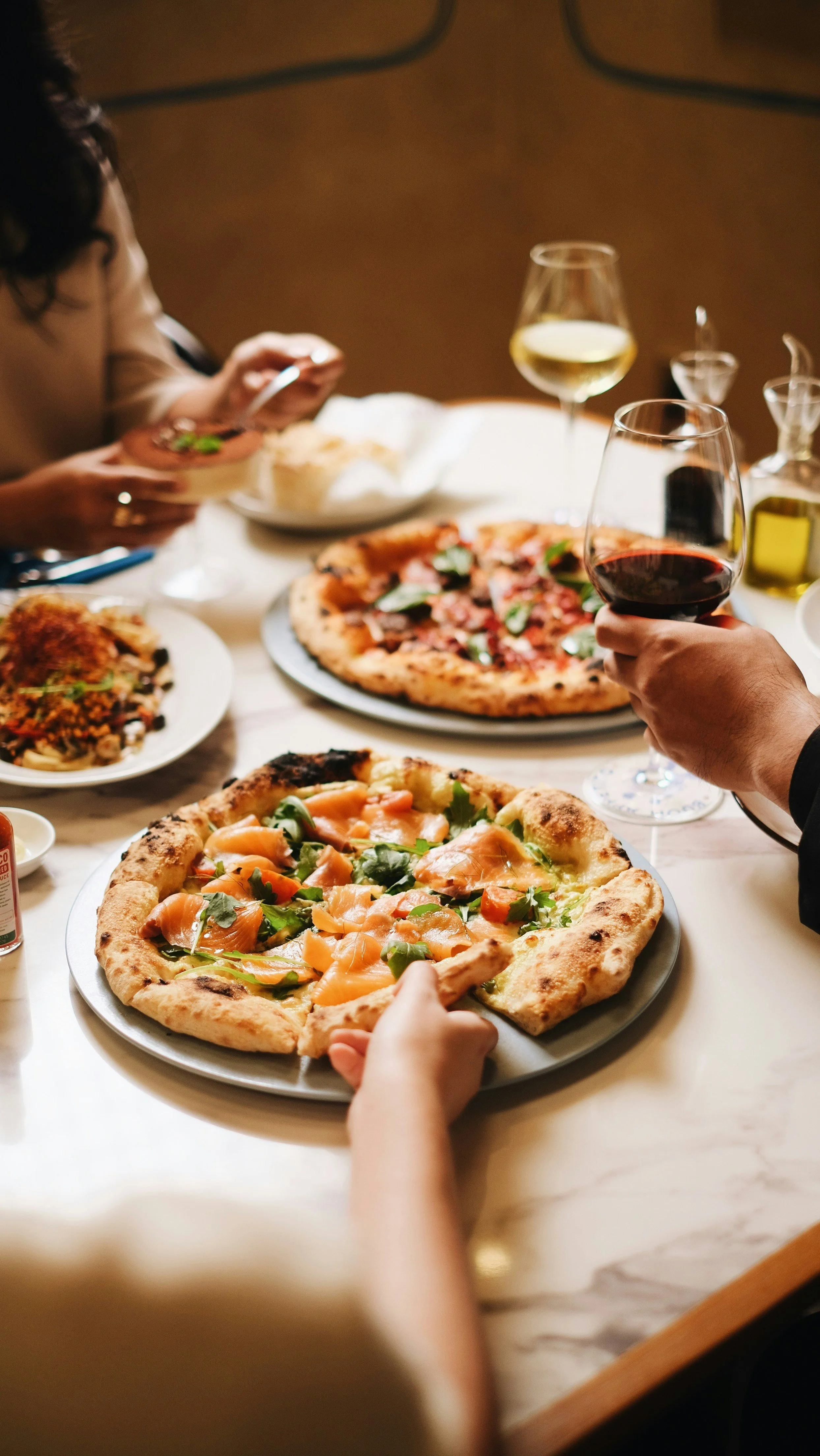 People enjoying a meal with two pizzas, glasses of white and red wine, and various dishes on a dining table.