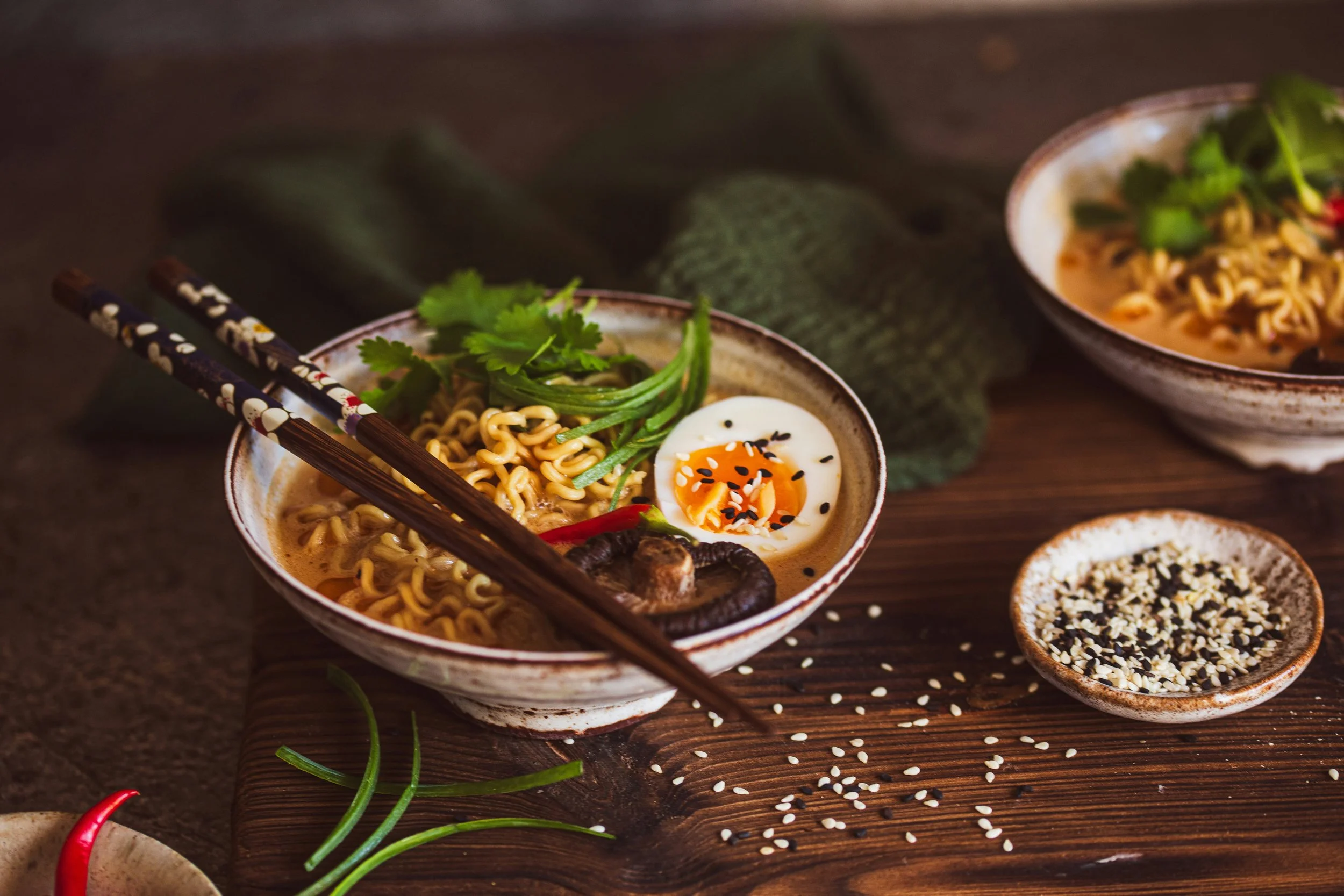 A bowl of ramen noodles garnished with herbs, a soft-boiled egg, and sesame seeds, with additional bowls of ramen and sesame seeds on the wooden table.