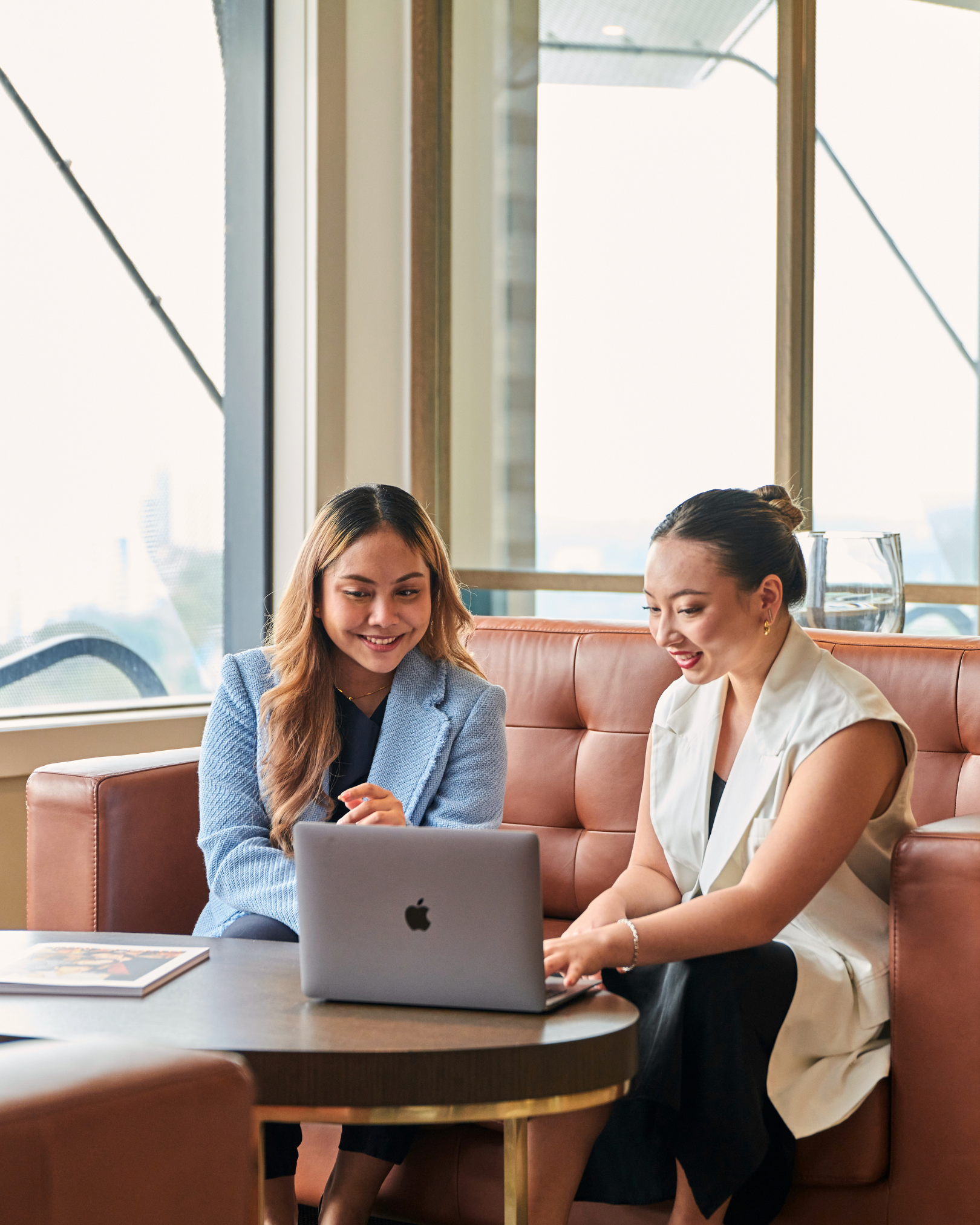 Two women sitting at a table in a brightly lit cafe, looking at a laptop together.