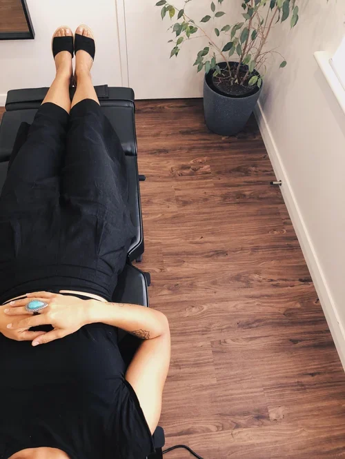 Person lying on a black chair with legs extended, wearing black clothing, a blue ring on their finger, and a tattoo on their arm. A potted plant sits on the wooden floor next to the chair.