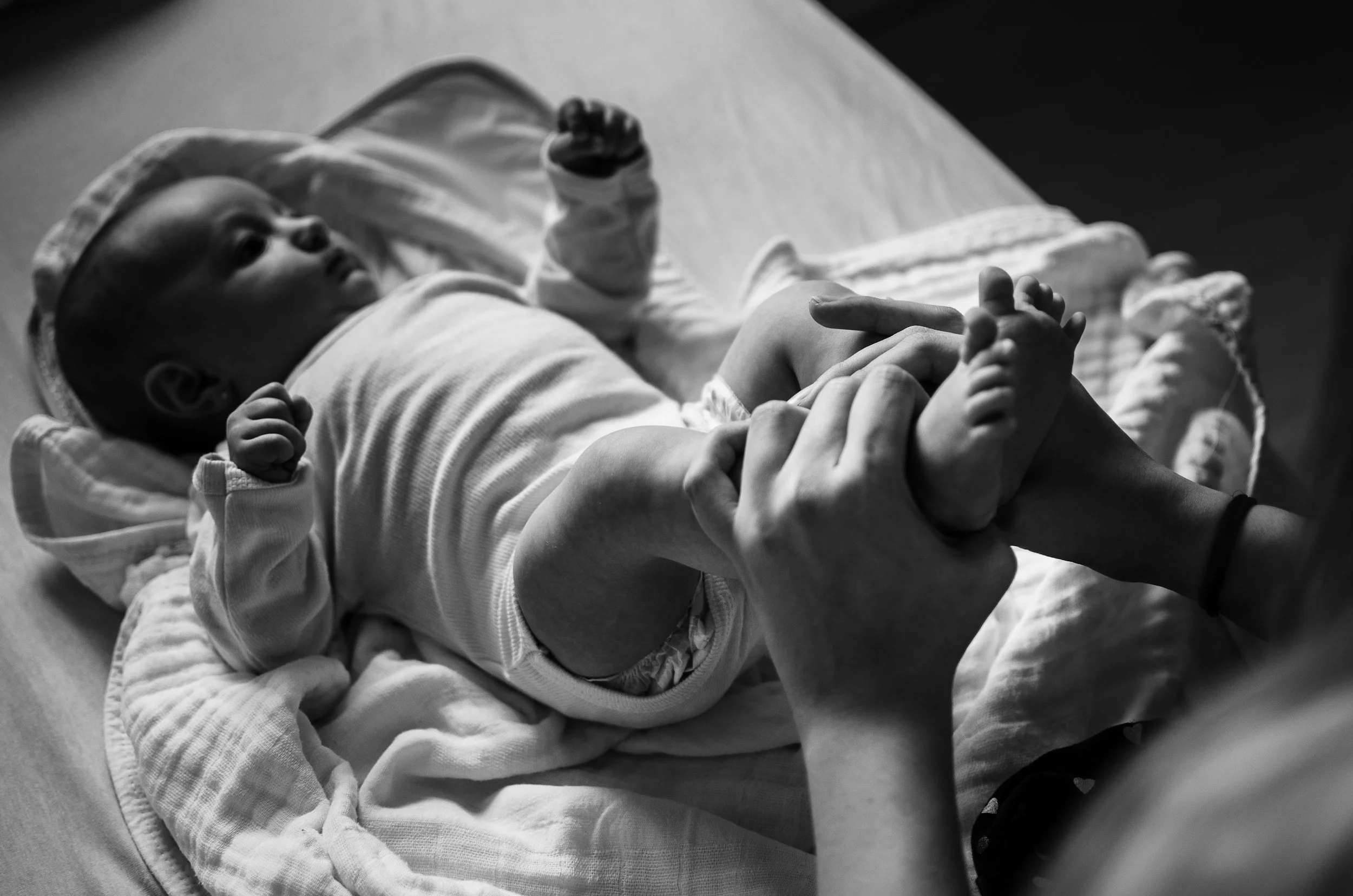 Black and white photo of a baby lying in a hospital bassinet, holding onto an adult's finger with both hands.