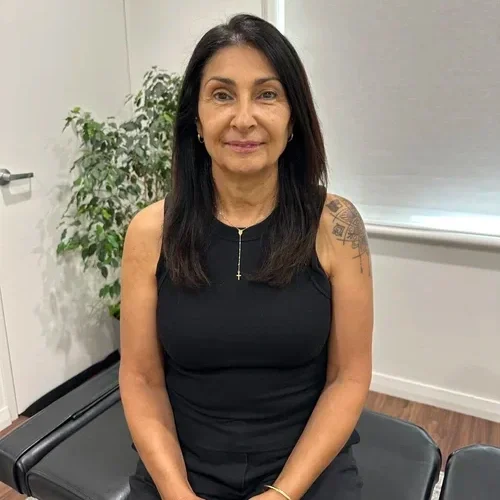 A woman with shoulder-length dark hair, wearing a black sleeveless top, sits on a chair in an indoor setting with a white wall, a closed door, and a plant in the background.