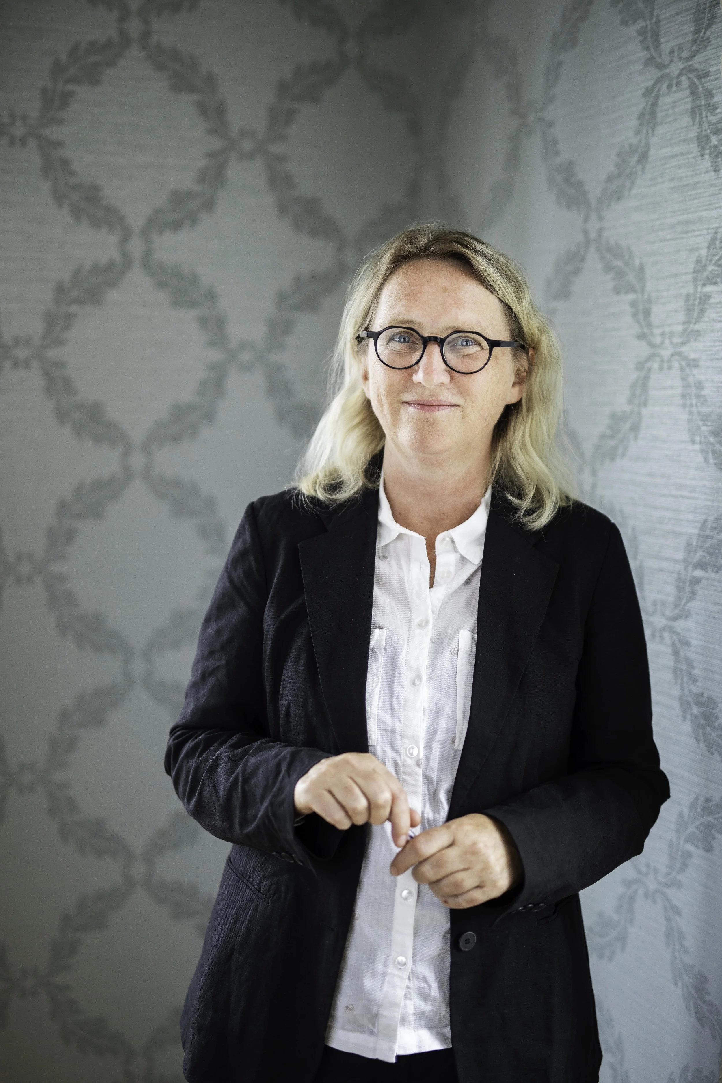En kvinne med blonde hår og svarte runde briller smiler. Hun er kledd i en svart blazer og hvit skjorte, og står foran en vegg med en dekorerende mønster.