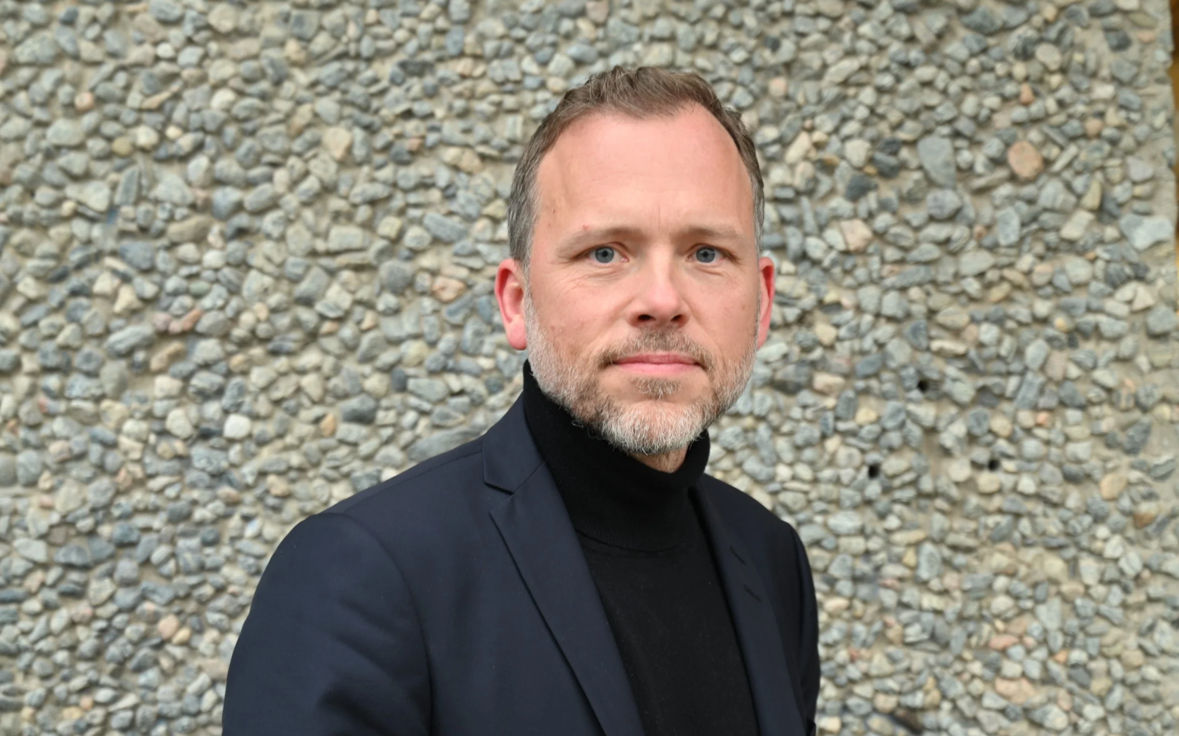 Mann i svart dress og turtleneck stående foran en steinvegg, ser inn i kamera.