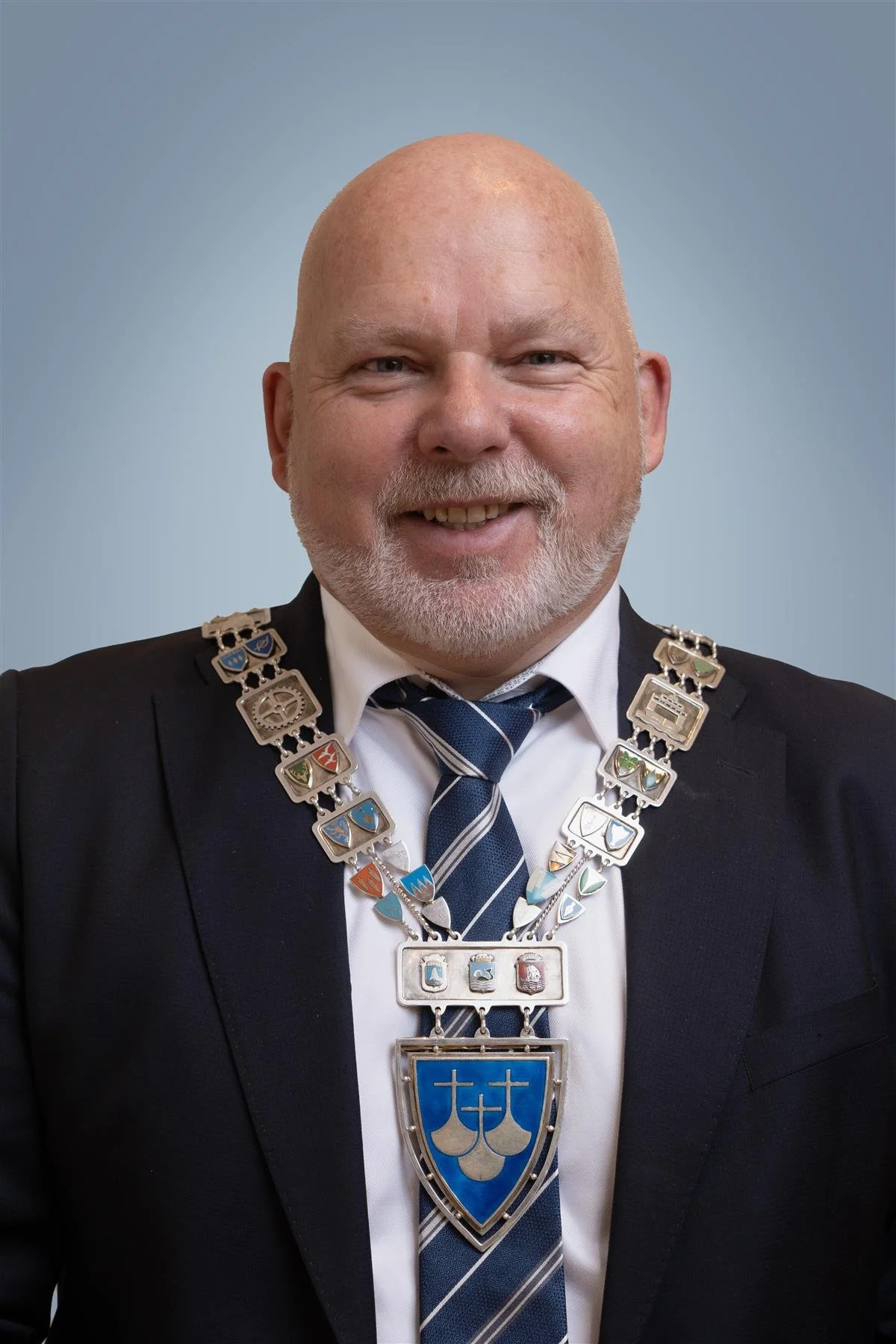 En mann med kort, lyst hår og skjegg smiler mot kamera. Han bærer en formell svart dress, hvit skjorte og en blå stripet slips. Han har på seg en stor medaljongkjede med flere små belter og et blått og sølv skjold med tre plussymboler.
