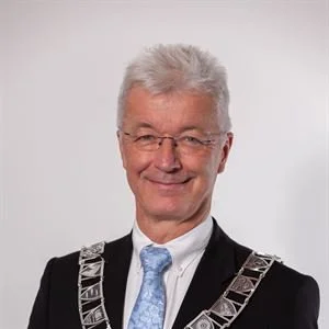 Portrett av en man iført dress og slips, med kort, grått hår og briller, smilende mot kameraet