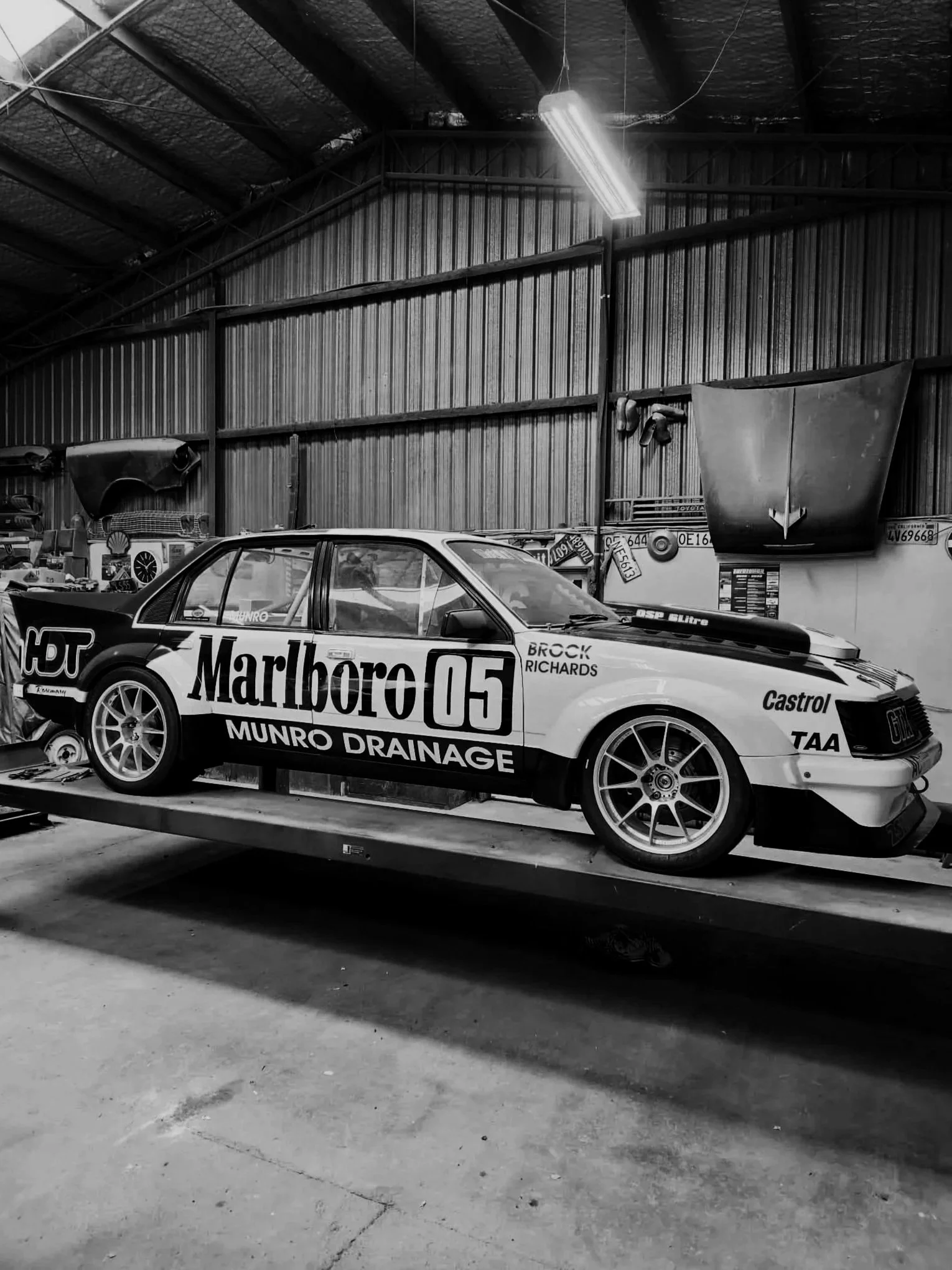 A race car with Marlboro branding and various sponsor logos, parked on a platform inside a garage or workshop with garage tools and parts in the background.