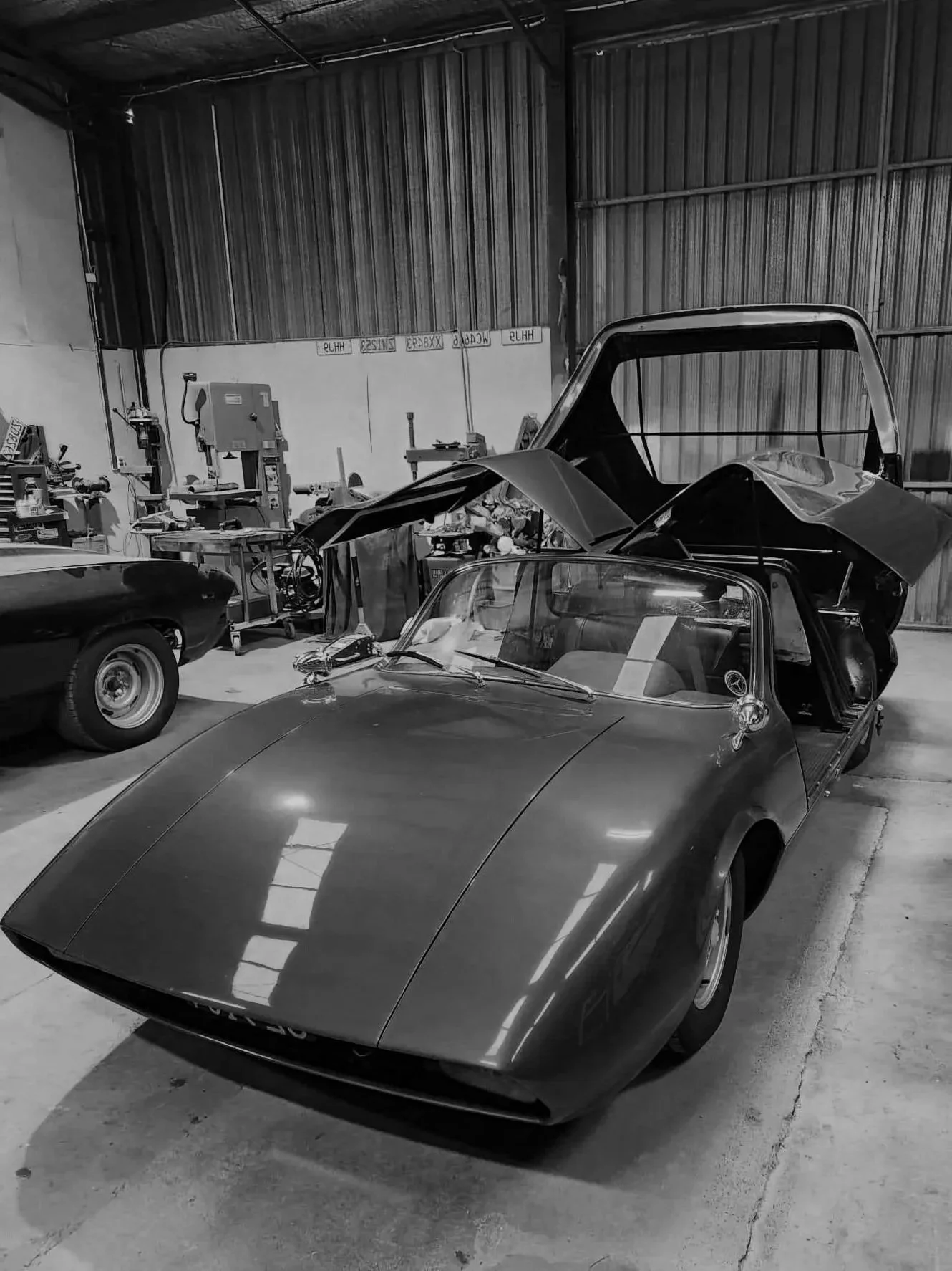 Black and white photo of a classic car with a sleek body and curved windshield, parked inside a garage with tools and equipment visible in the background.