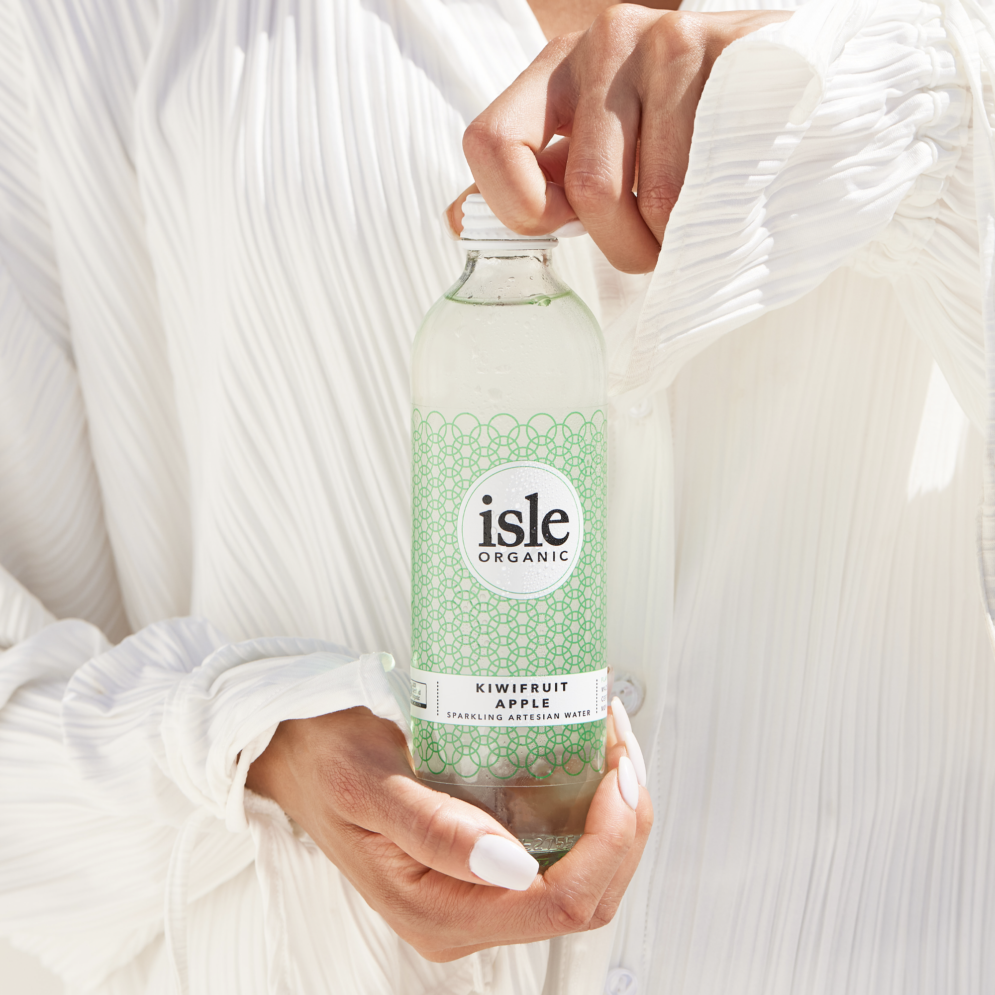 A woman holding a glass bottle of isle organic sparkling artesian spring water, wearing with a creamy white, textured linen top.