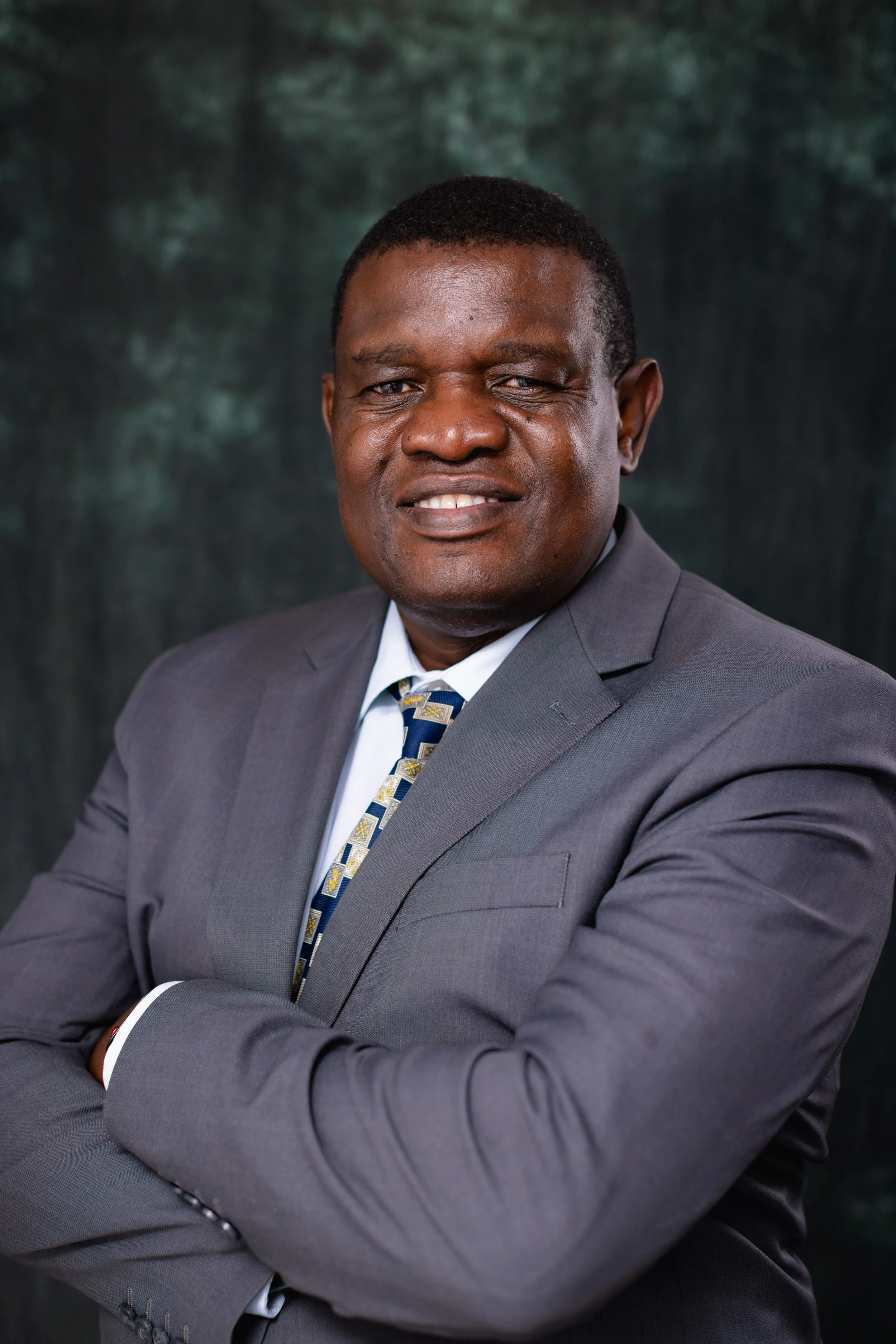 Portrait of an African American man in a gray suit and patterned tie, smiling slightly, with arms crossed, against a dark blurred background.