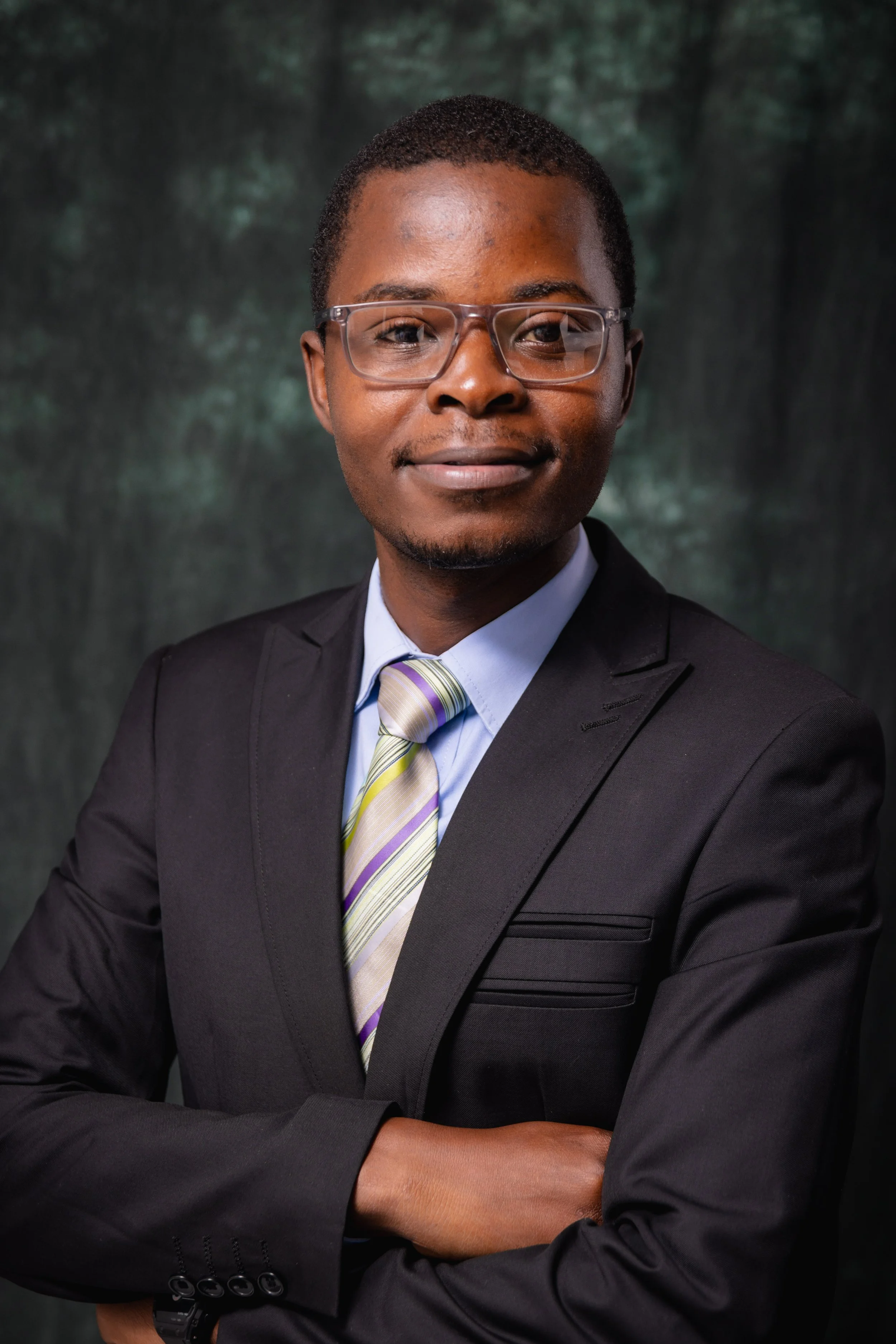 A person wearing glasses, a dark suit, a white shirt, and a multicolored striped tie, with arms crossed, in front of a green forest background.