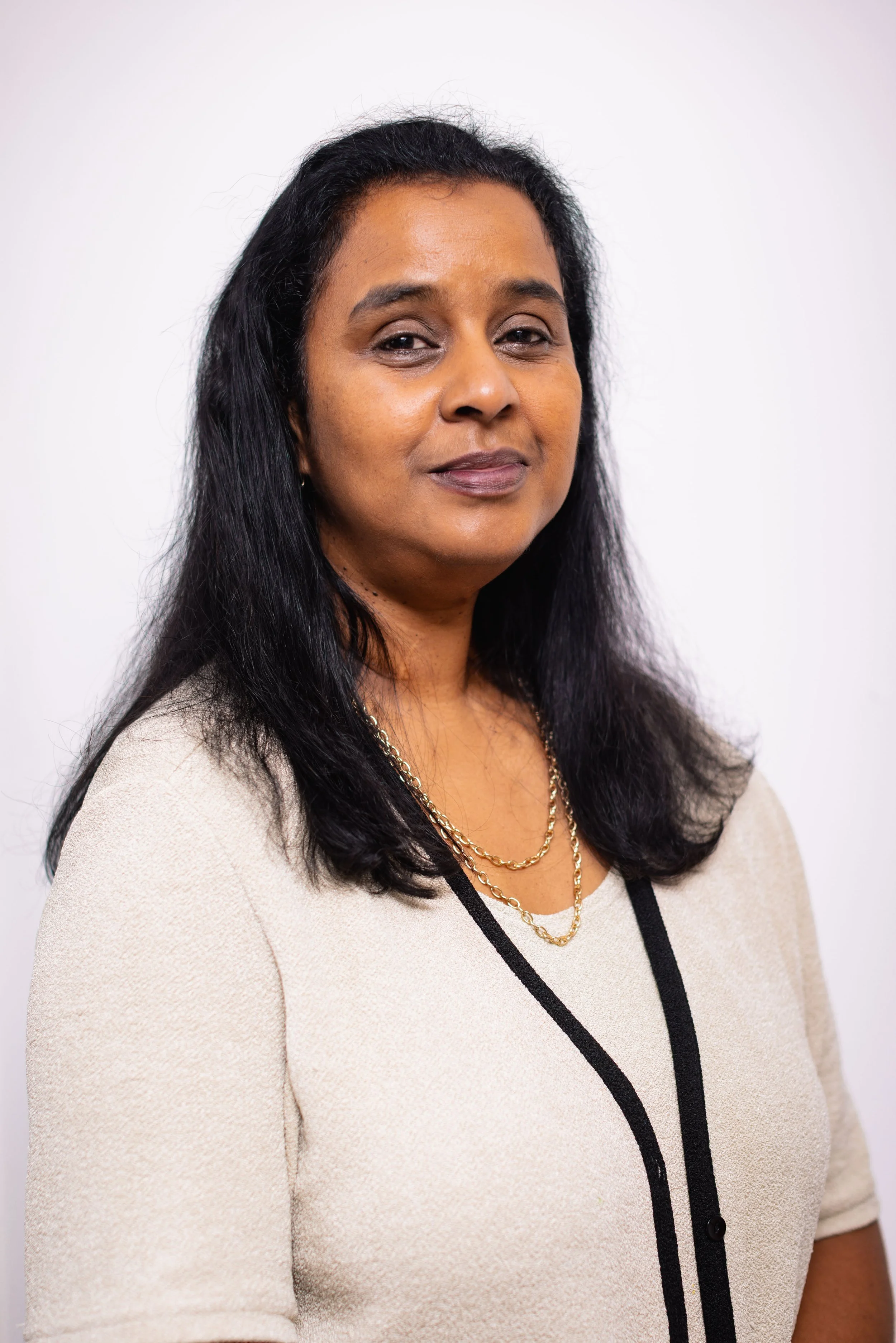 A woman with long black hair, wearing a beige sweater with black trim and gold necklaces, standing against a white background.