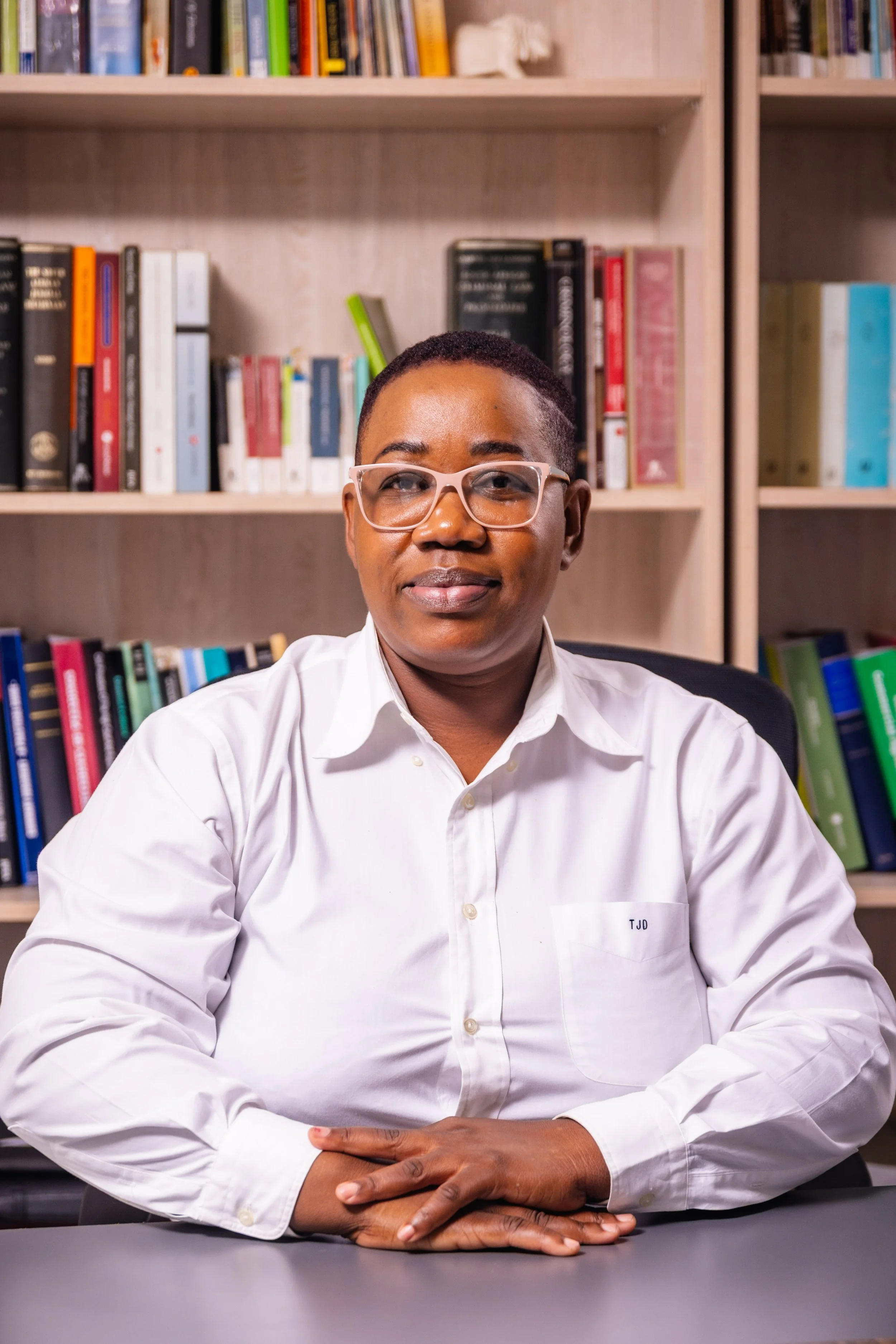 A person with short hair, wearing glasses and a white collared shirt, sitting at a desk in front of a bookshelf filled with books.