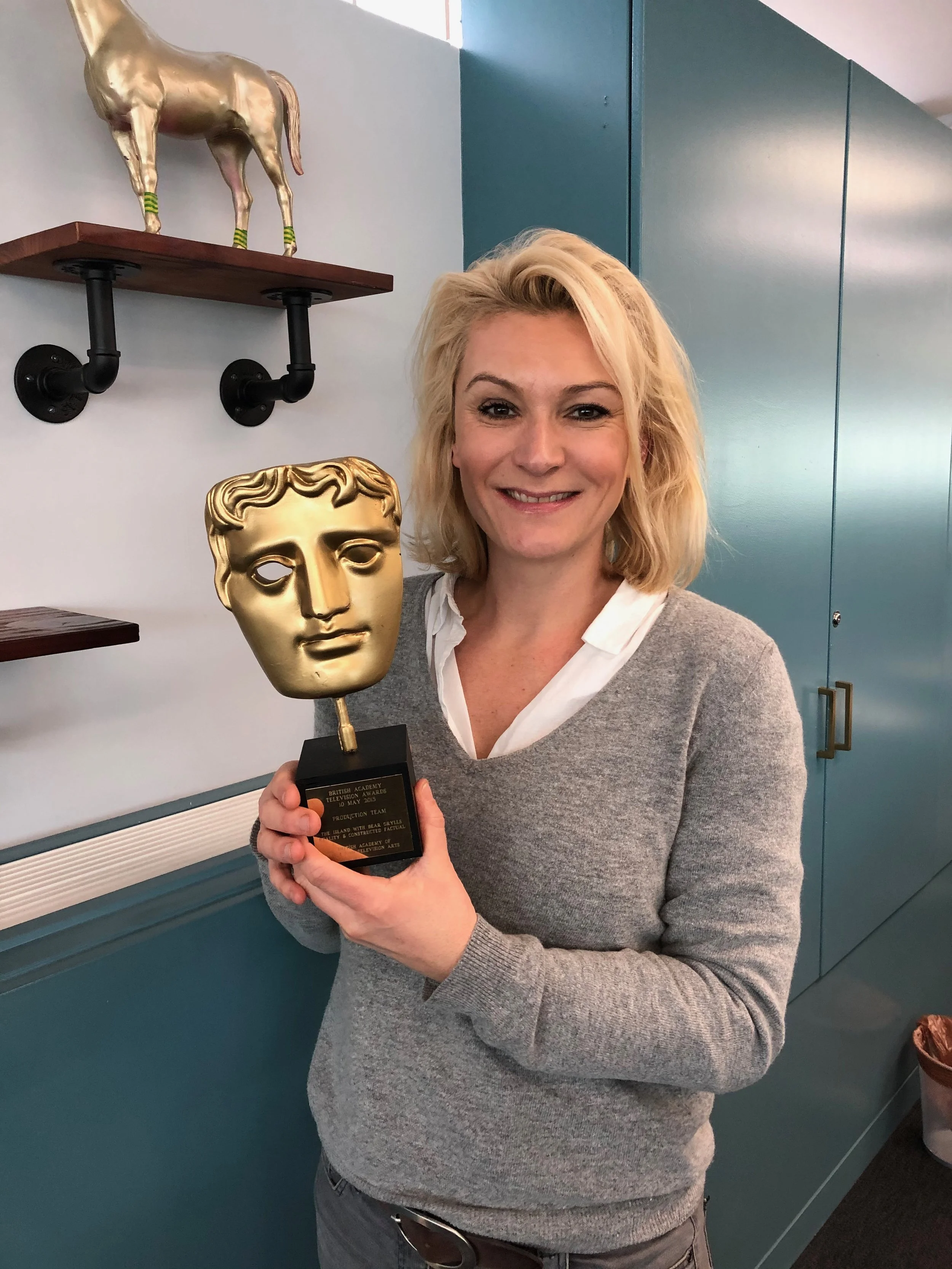 Woman with blonde hair smiling while holding a BAFTA award, standing in front of blue lockers.