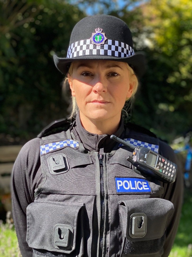 A female police officer outdoors wearing a black uniform and hat with a checkered band, featuring a police badge and radio, with trees and foliage in the background.