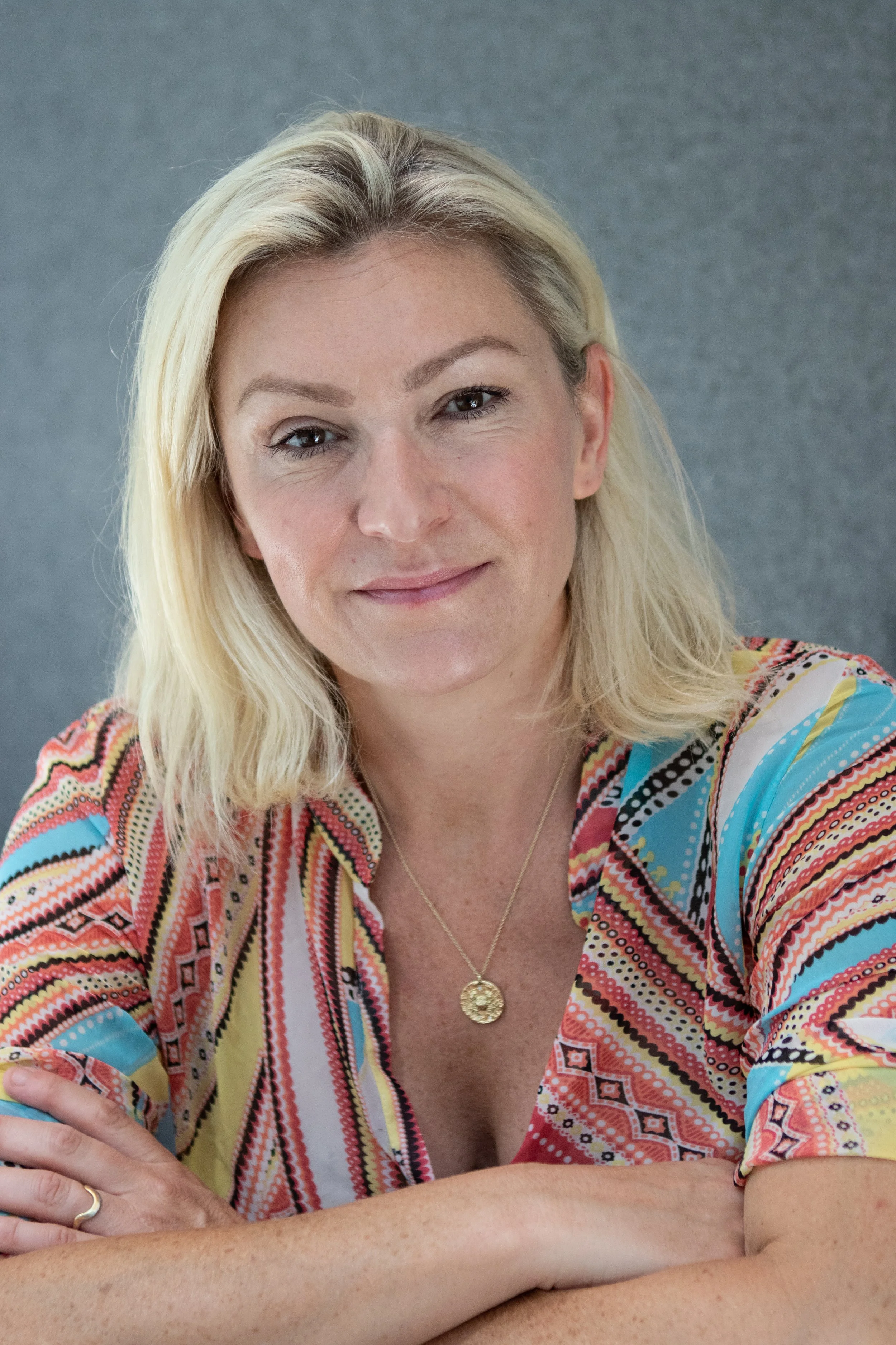 Close-up portrait of a smiling blonde woman with wavy hair, wearing a colorful patterned blouse and a gold pendant necklace, posing in front of a gray background.
