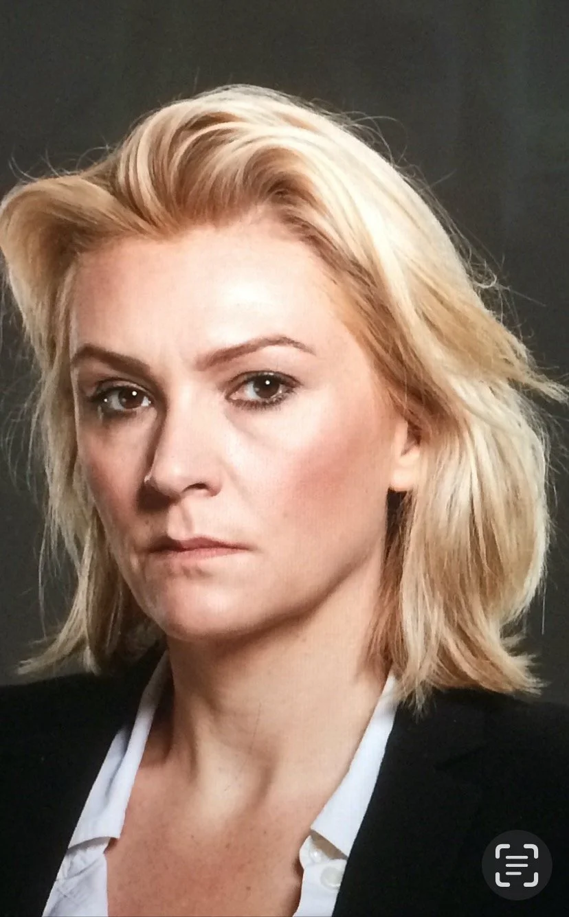 Close-up of a woman with blonde hair and a serious expression, wearing a white shirt and black blazer against a dark background.